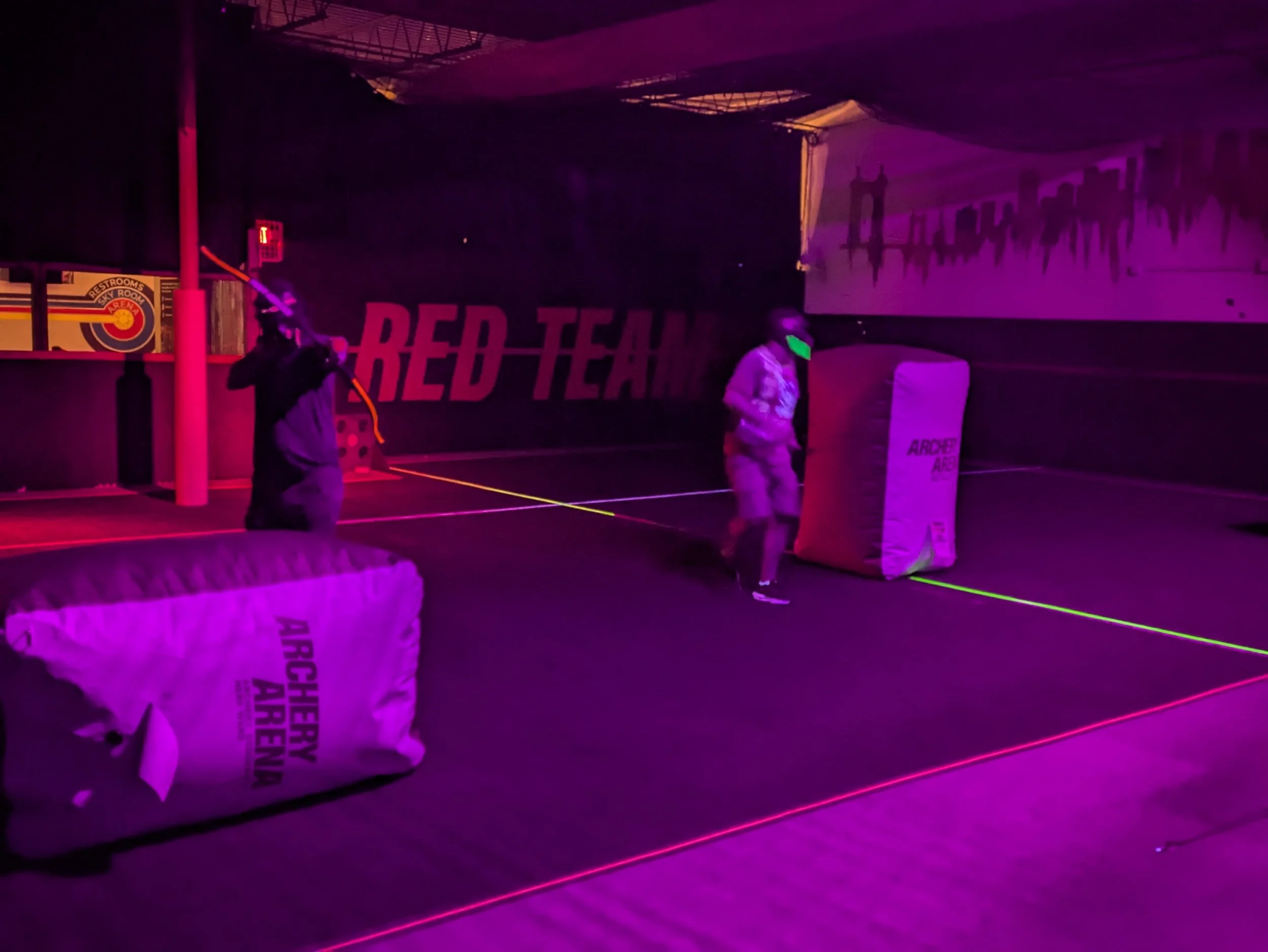 People playing laser tag in a dark room with neon lights, target barriers, and the sign 'Red Team' on the wall.