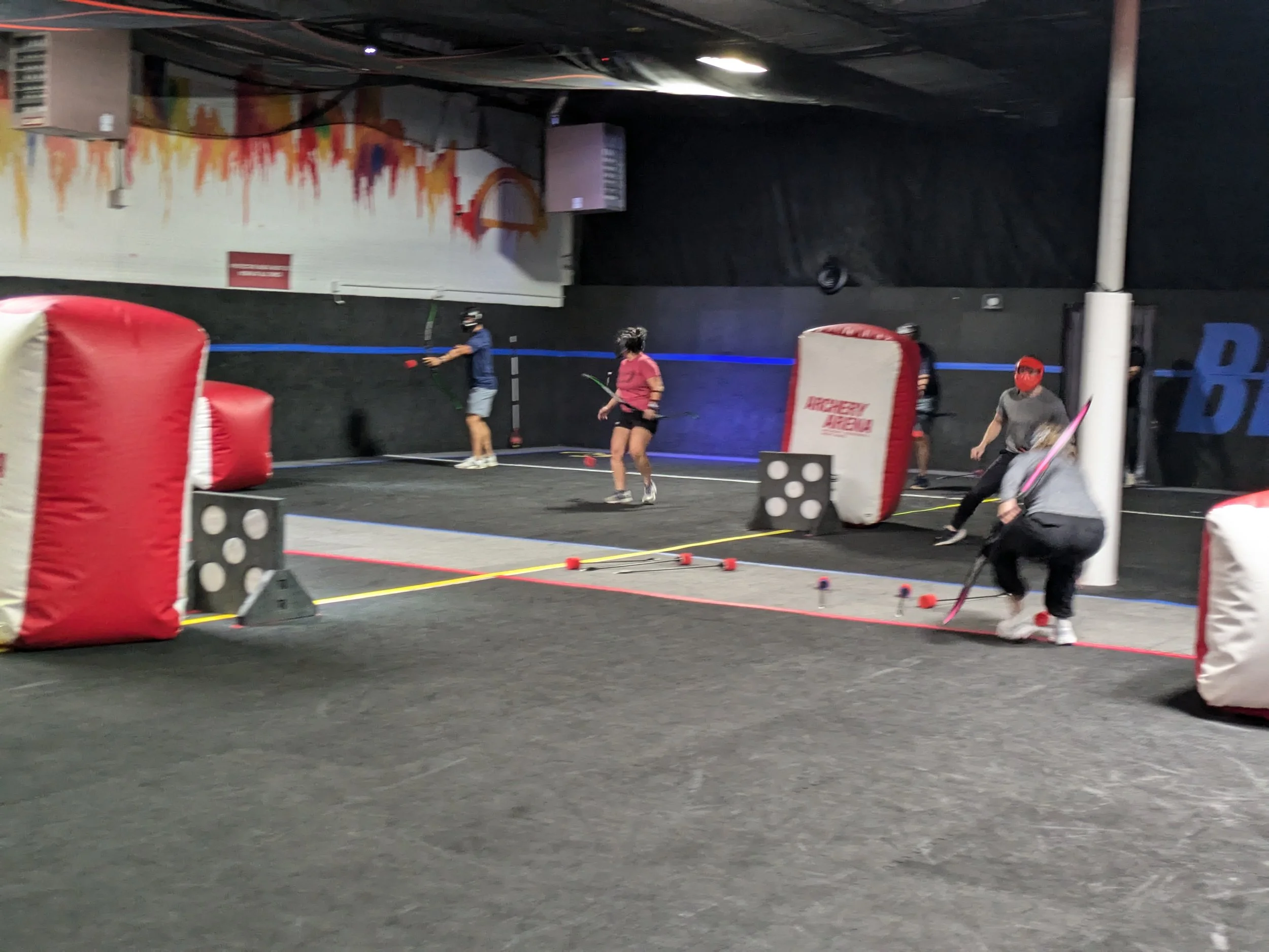Indoor laser tag arena with players wearing masks and holding laser guns, behind large foam barriers, on a dark floor with colored lines and scattered foam balls.