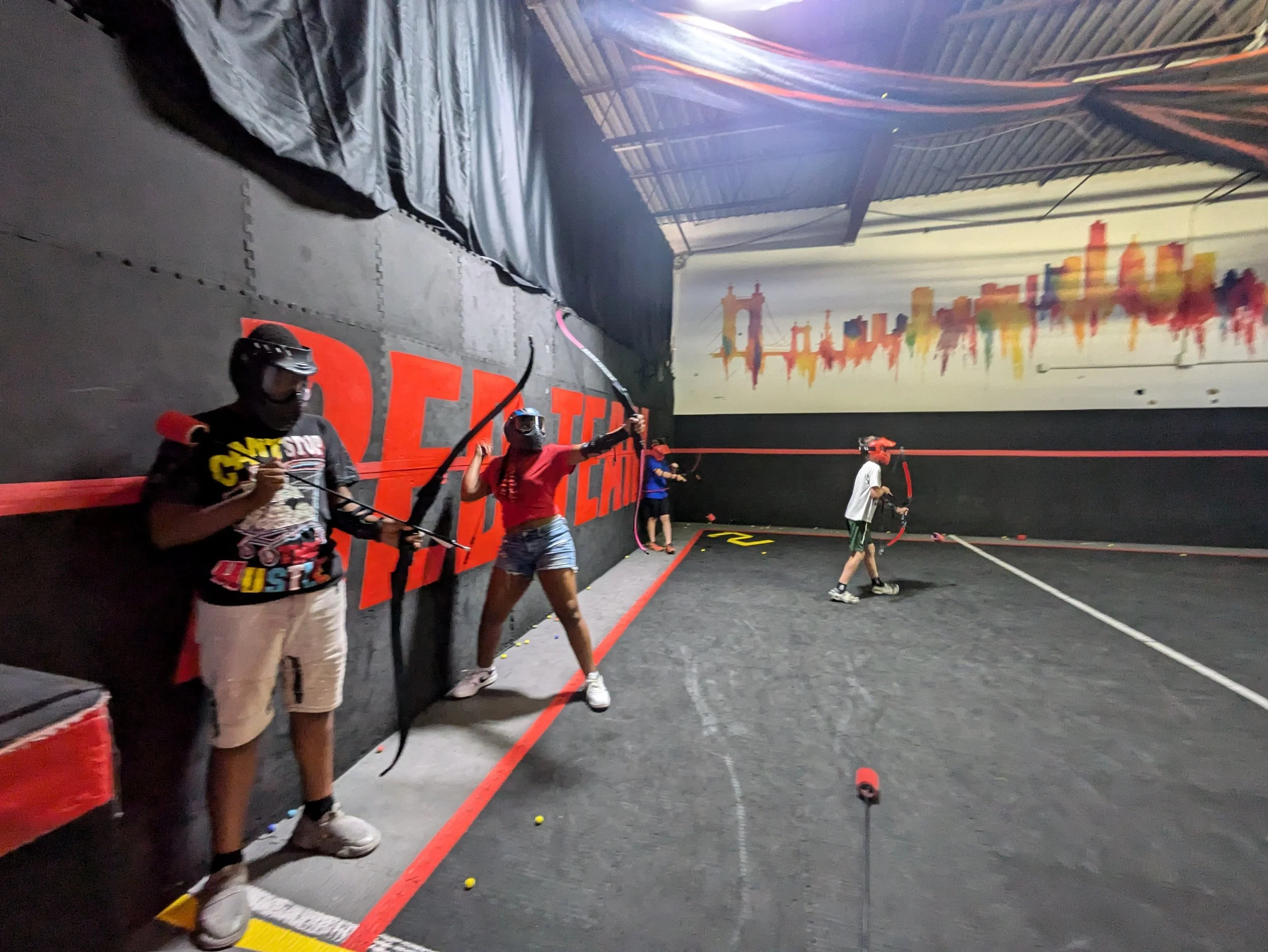 Kids and teens wearing helmets and protective gear playing indoor archery tag, holding bows and arrows inside a black-walled gym with colorful cityscape artwork on the wall.