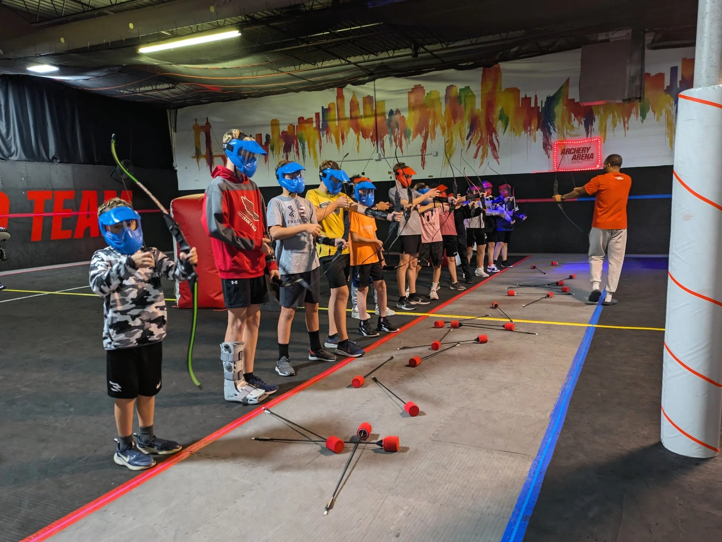 Children and a coach practicing archery indoors, all wearing blue masks, with arrows on the ground, and a colorful cityscape mural on the wall.