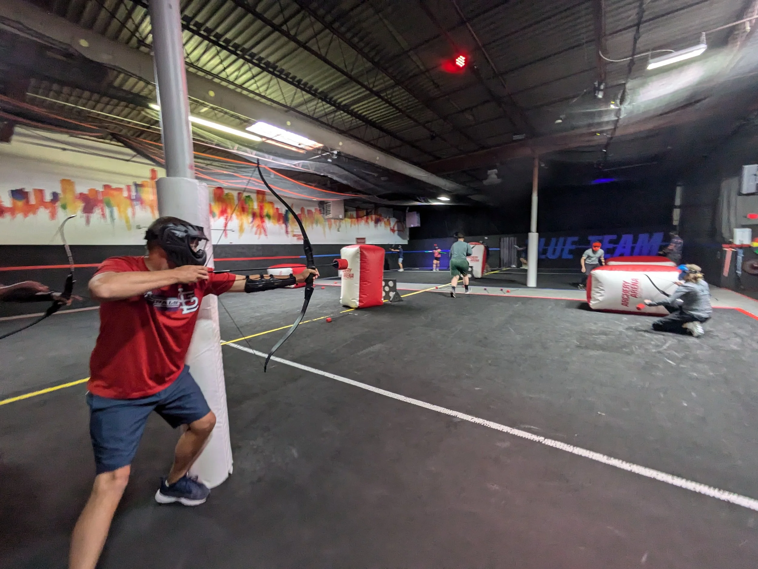 People playing an indoor archery tag game, shooting foam-tipped arrows in a dimly lit space with colorful wall art and inflatable barriers.