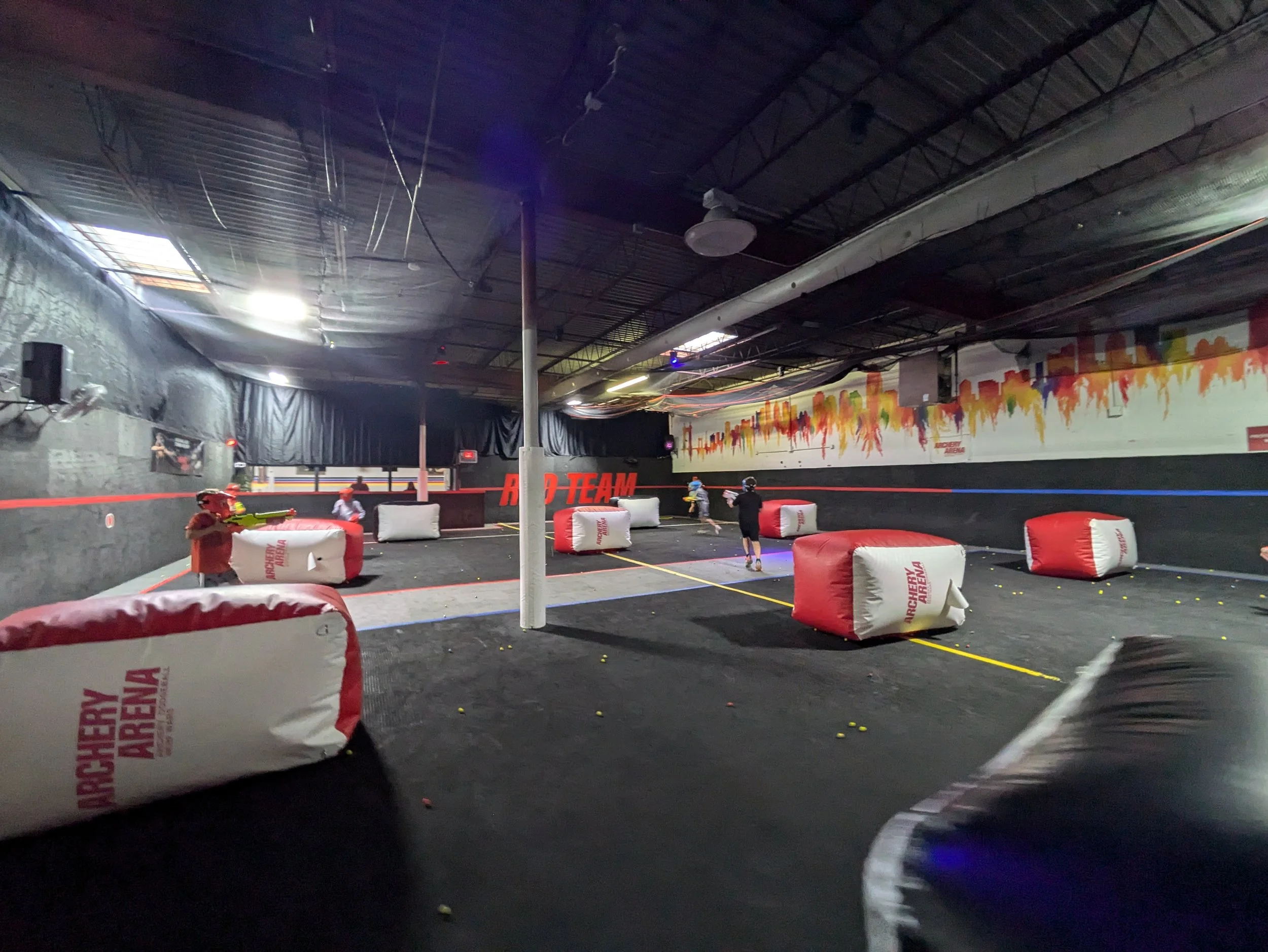 Indoor laser tag arena with large inflatable cover objects, players with laser guns, black walls, and colorful graffiti-style artwork on wall.