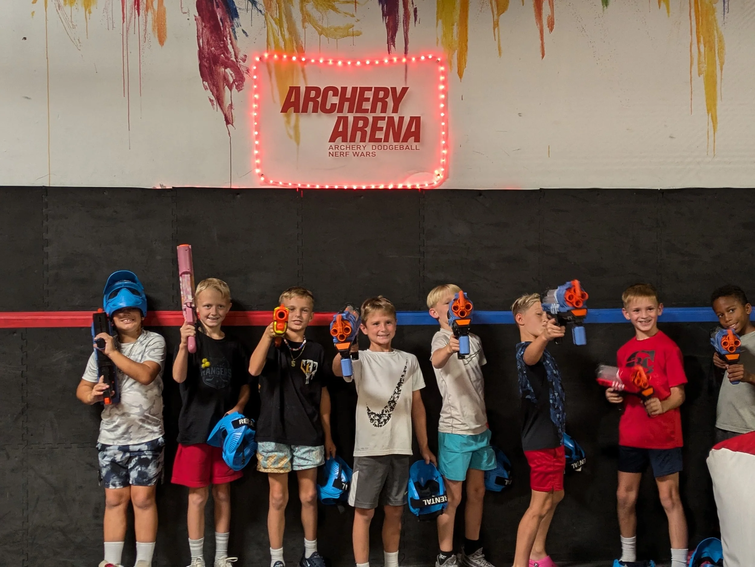 Kids holding toy guns at an indoor nerf gun arena, standing in front of a sign that reads 'Archery Arena' with neon lights.