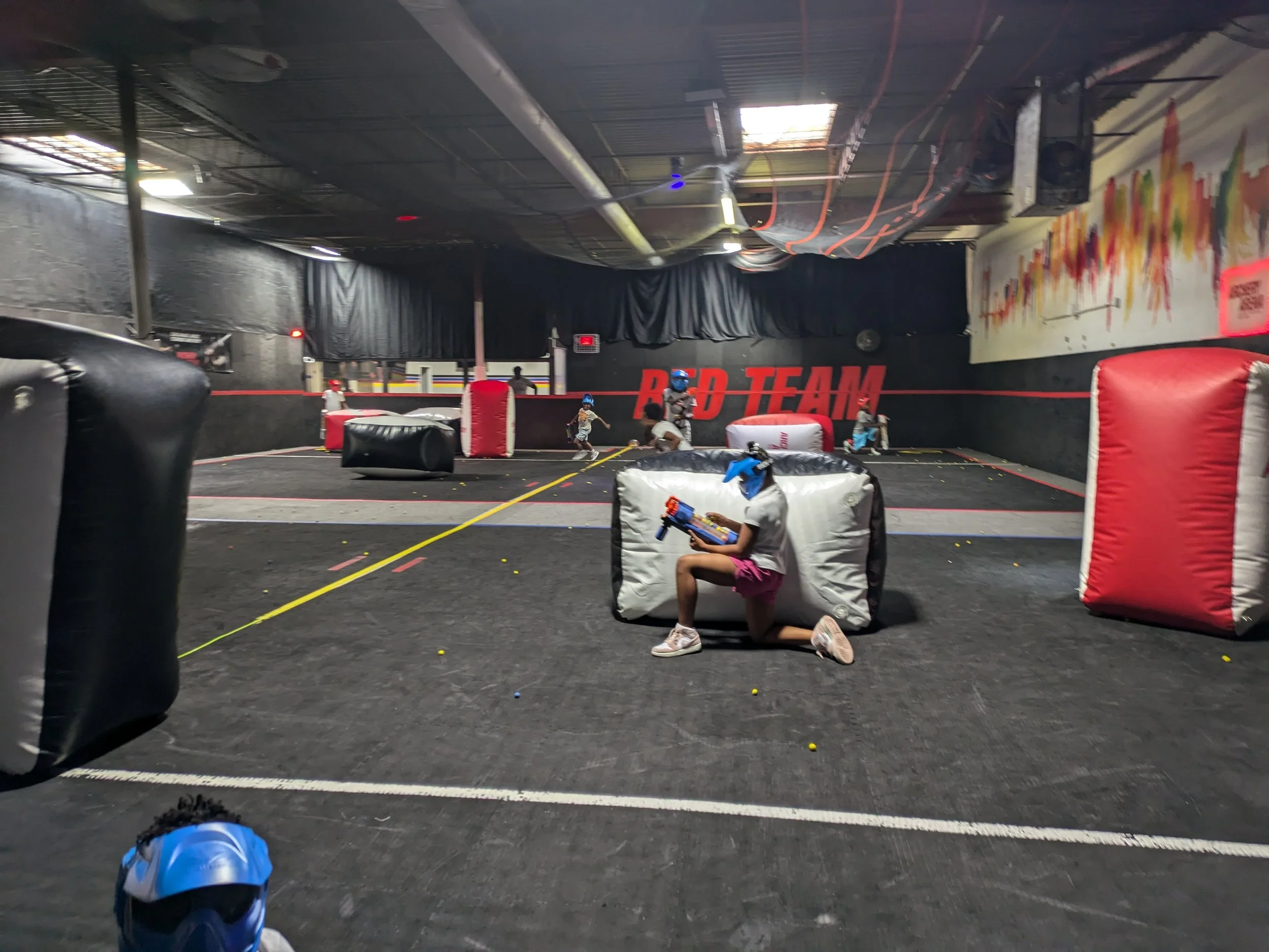 People playing indoor laser tag or paintball game with protective gear and foam weapons in an enclosed arena with black walls, some with colorful graffiti, and obstacles.