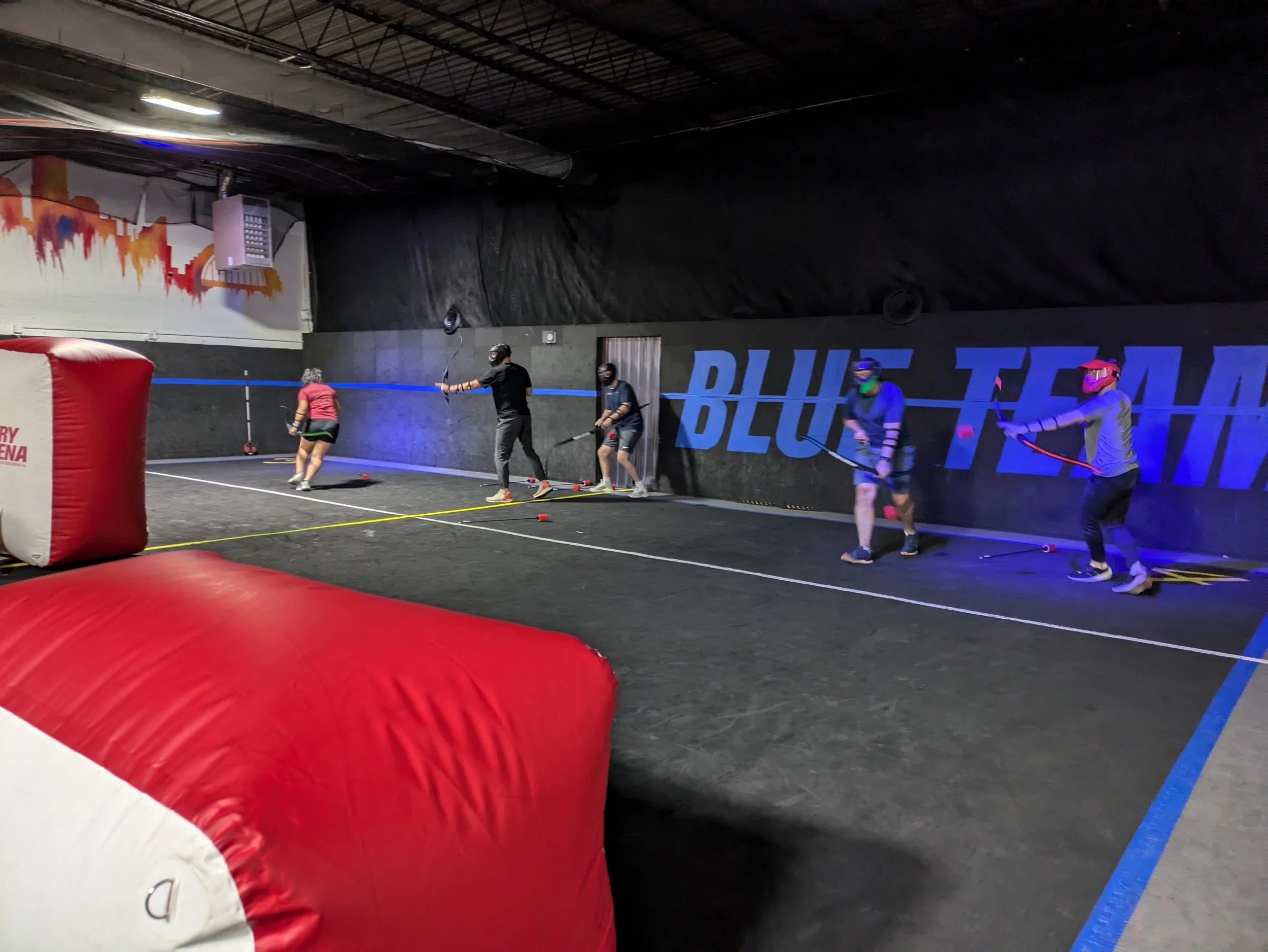 Indoor hockey arena with six players practicing hockey sticks. The wall displays large blue text 'BLUE' on black background. Large red and white inflatable bumper in the foreground.