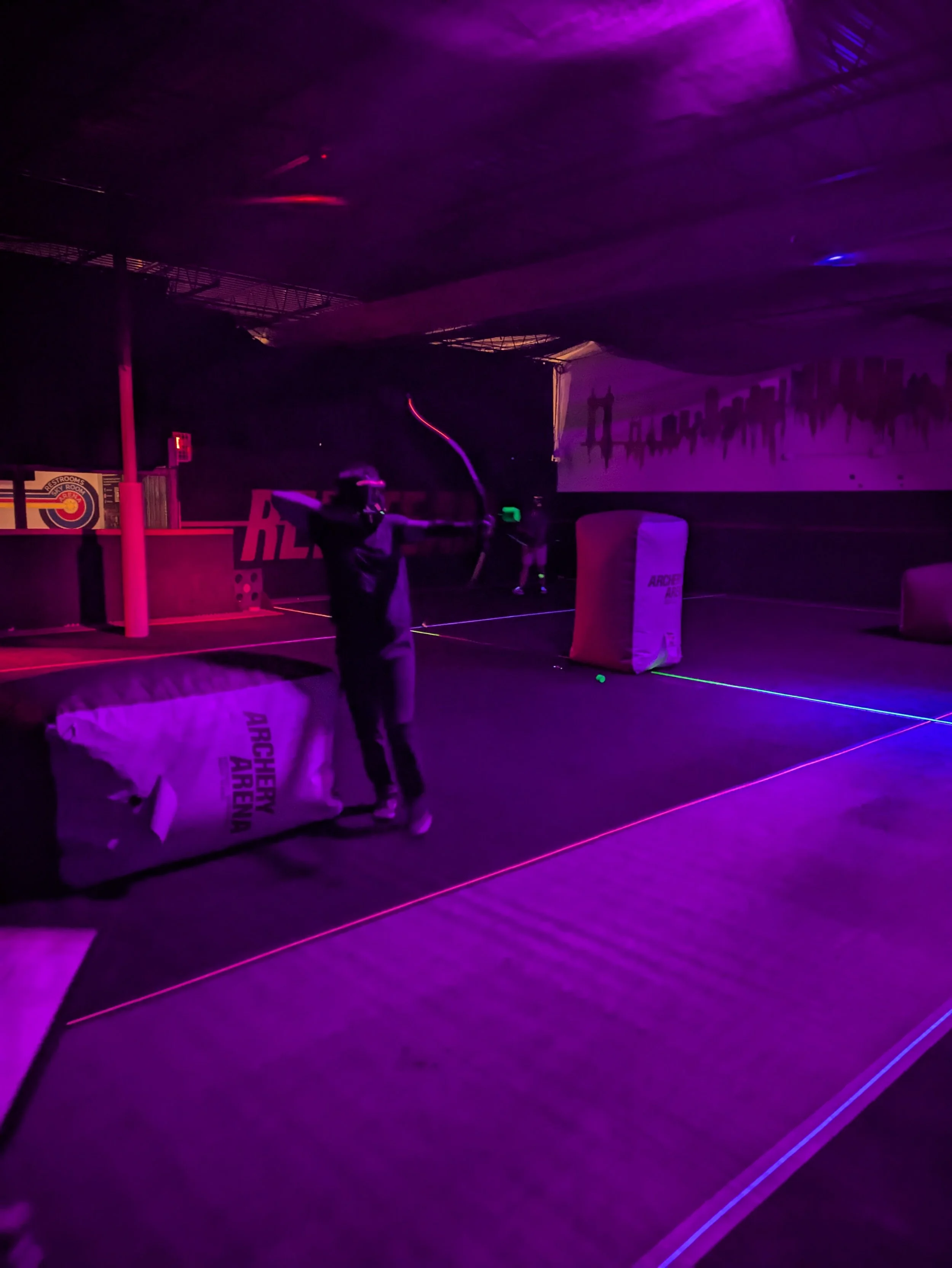 Person playing archery tag in a dark indoor arena with neon lighting, holding a bow and arrow, and wearing a laser tag vest. The arena has inflatable barriers and neon lines on the floor.