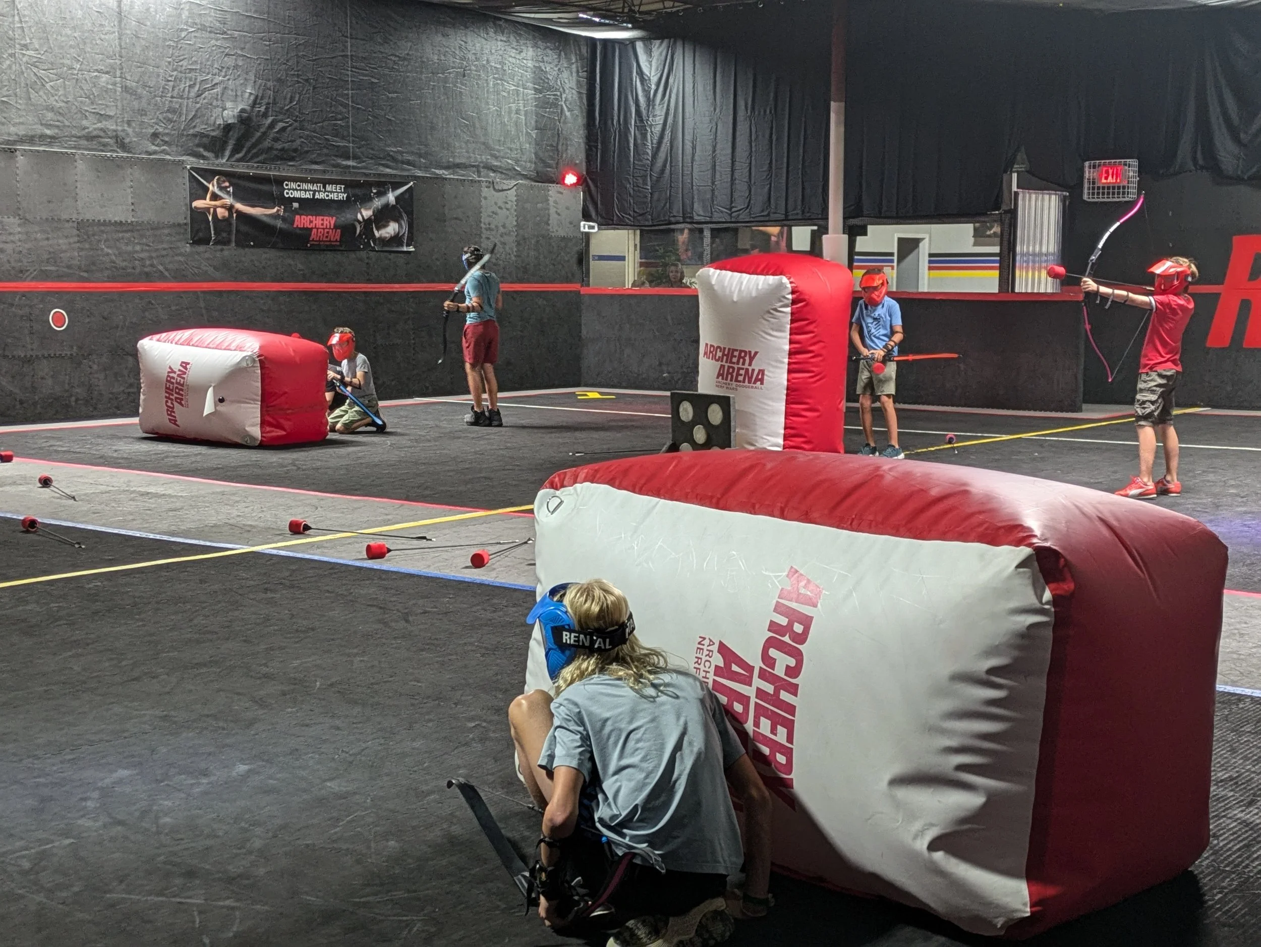 Children practicing archery indoors behind inflatable barriers, with adults supervising, in a training facility at Archery Arena.