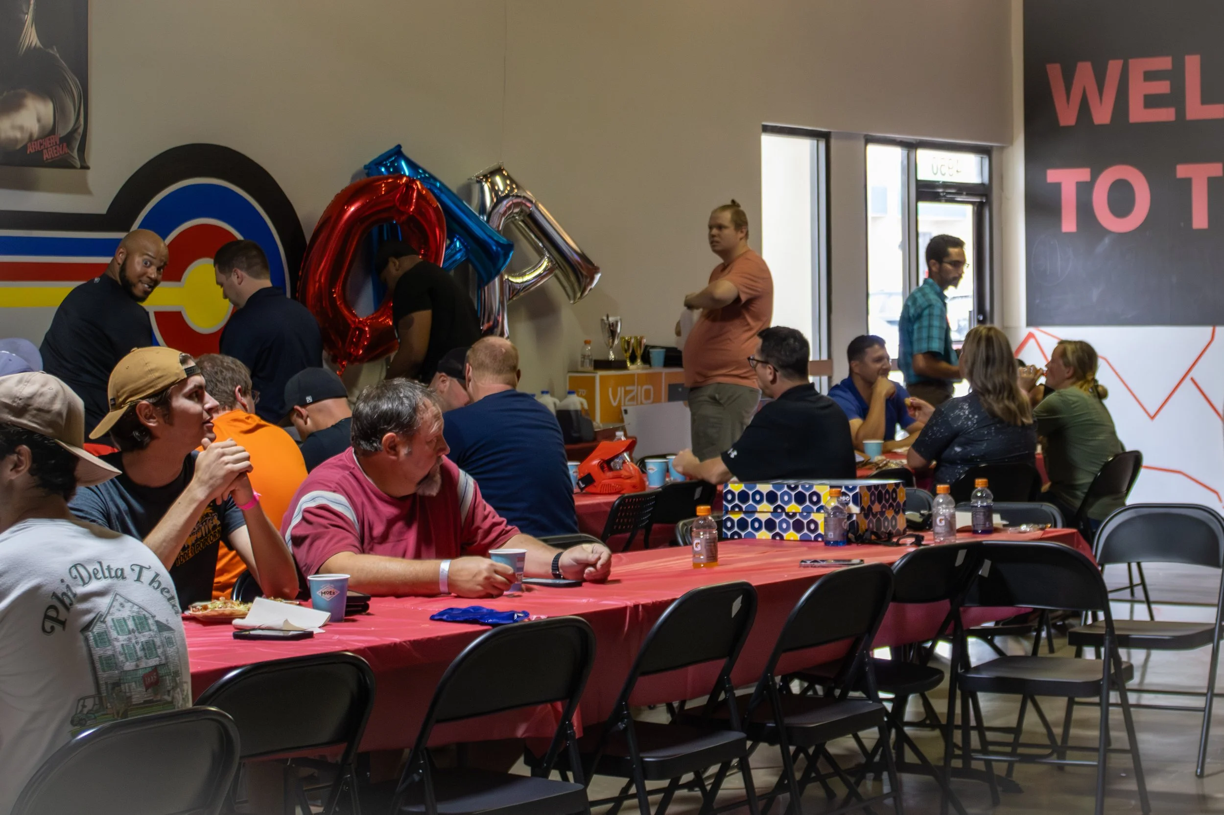 People gathered at a room with tables, celebrating with balloons and snacks, some sitting and talking, others standing.