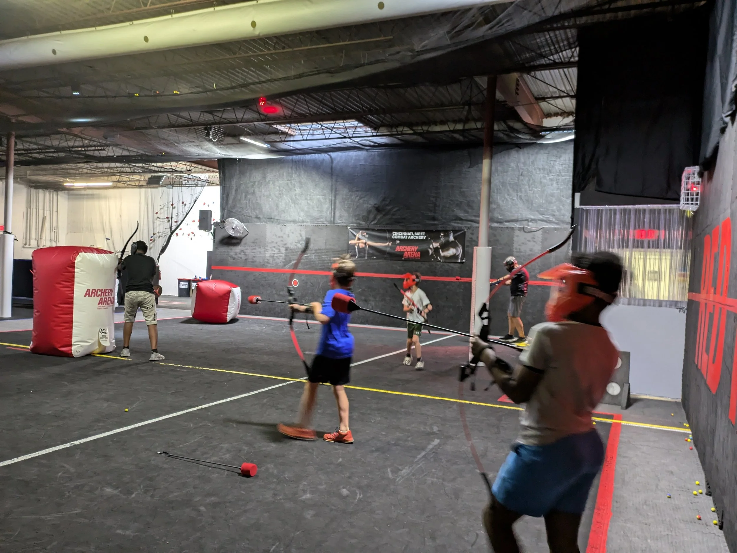 Indoor archery range with children and adults practicing archery using bows and safety gear, targets, and padded barriers.