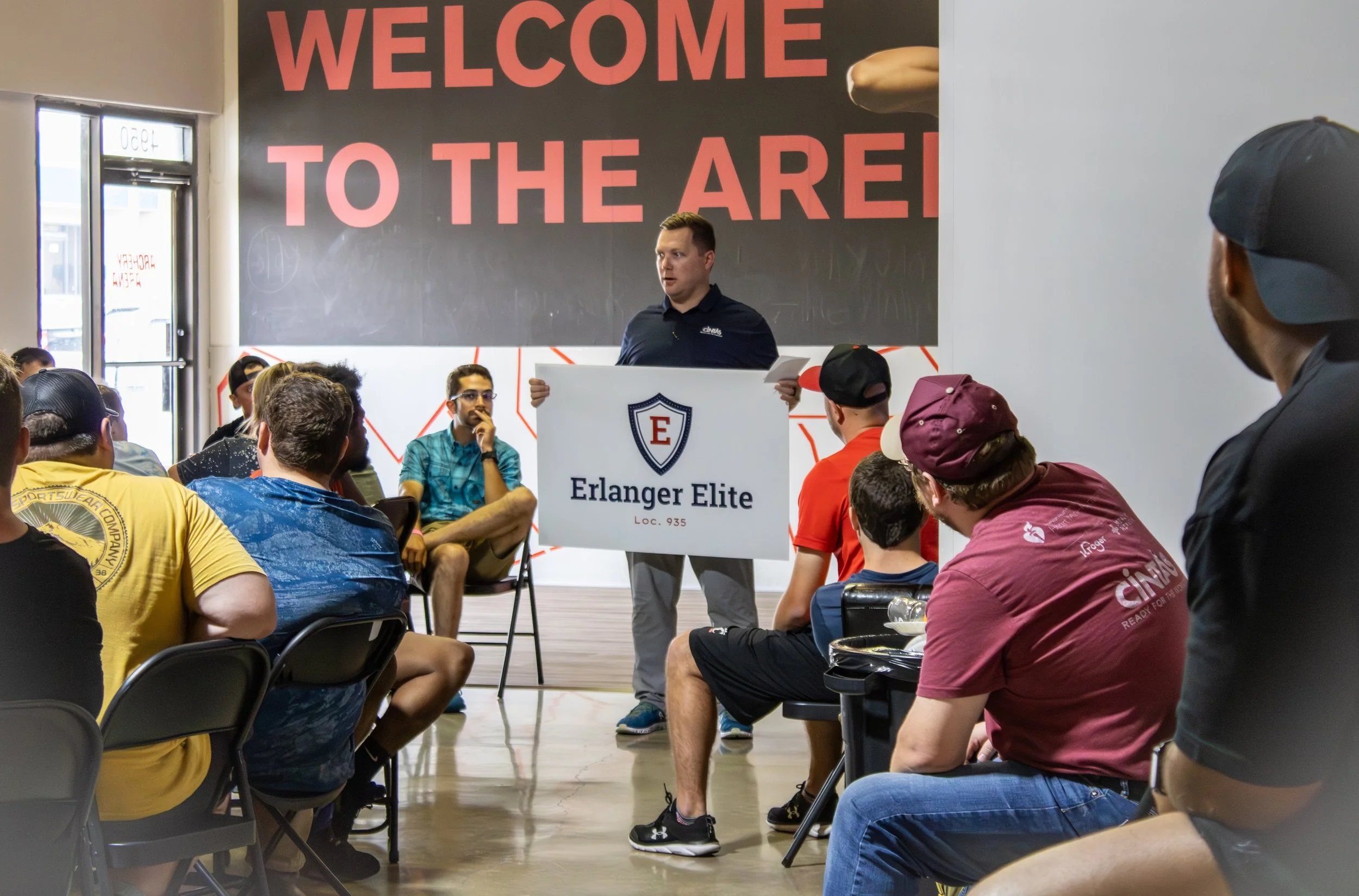 A speaker holds a sign reading 'Erlanger Elite' as he addresses an audience sitting in chairs inside a gym or event space with a large sign behind him that says 'Welcome to the Arena' in bold red and pink letters.