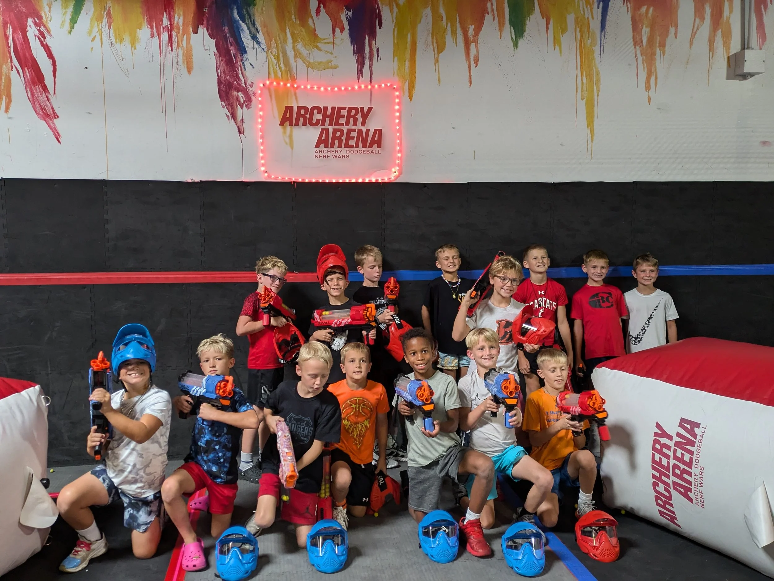 Children at an indoor laser tag arena posing with laser guns and wearing protective helmets, with a sign that reads 'Archery Arena' in the background.