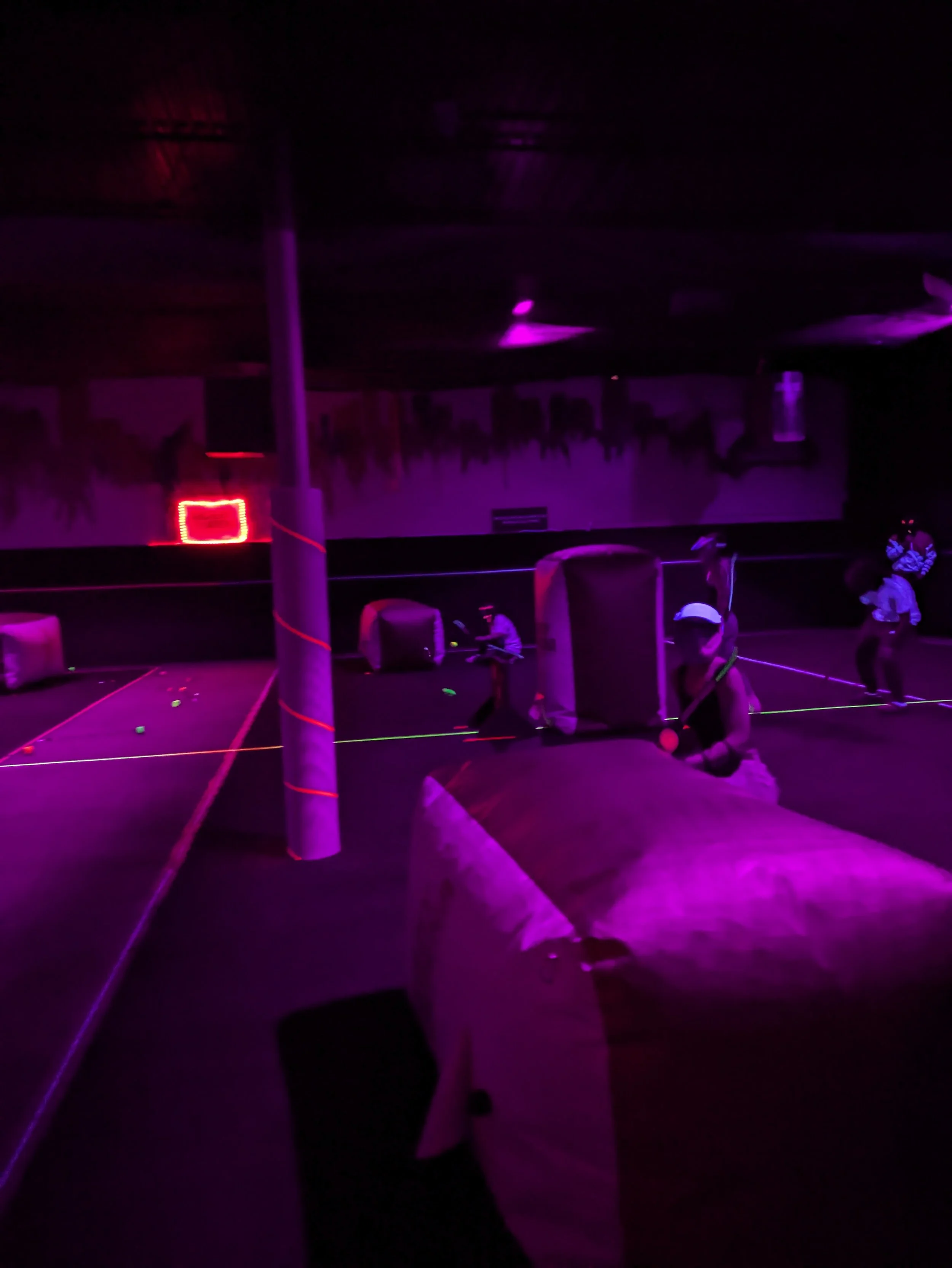 Indoor laser tag arena with players wearing vests and helmets, illuminated by black lights and neon colors.