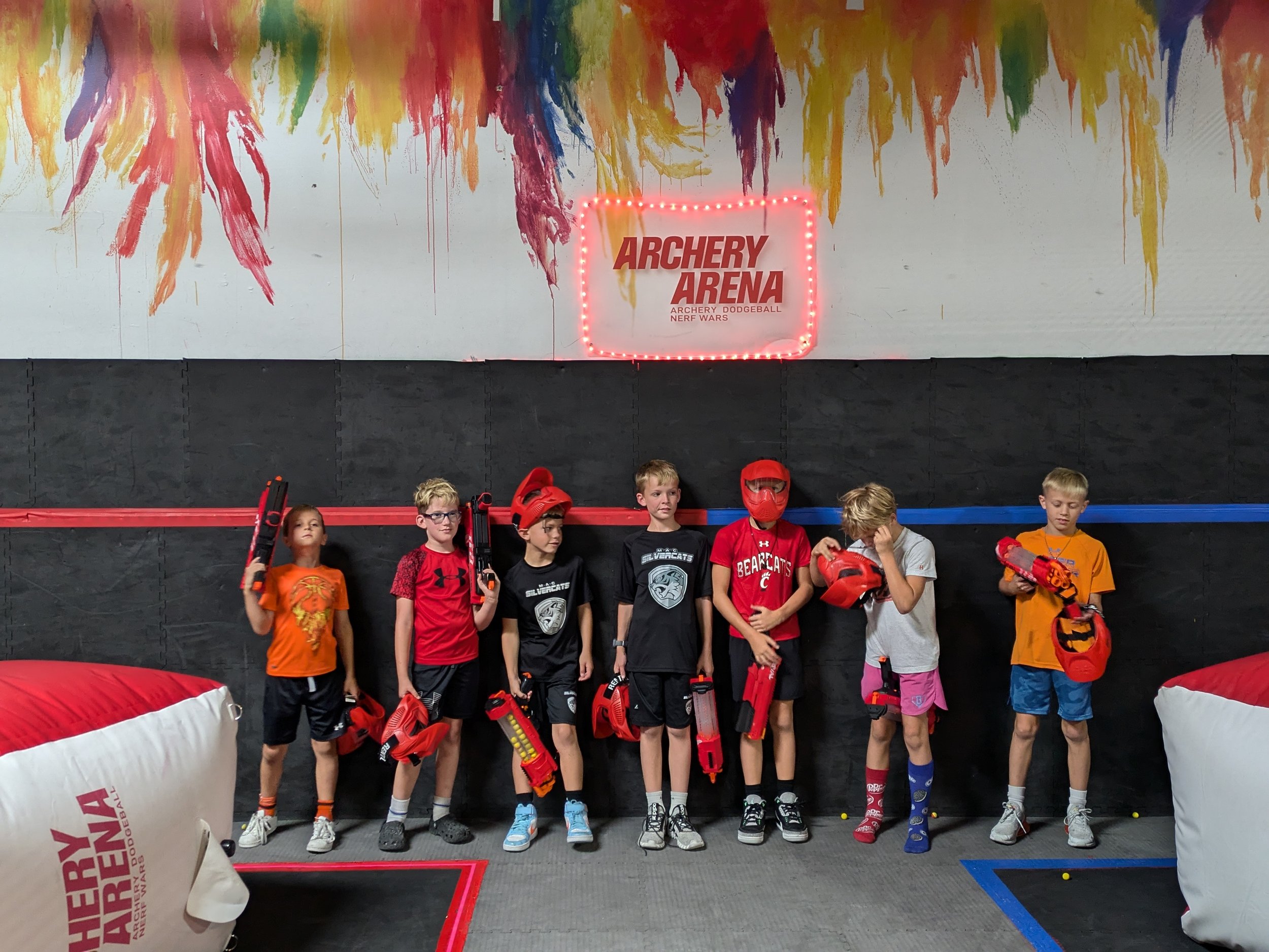 Group of seven children standing in line at archery arena with foam weapons, some with protective masks and gear, colorful wall art, and neon sign reading 'Archery Arena'.