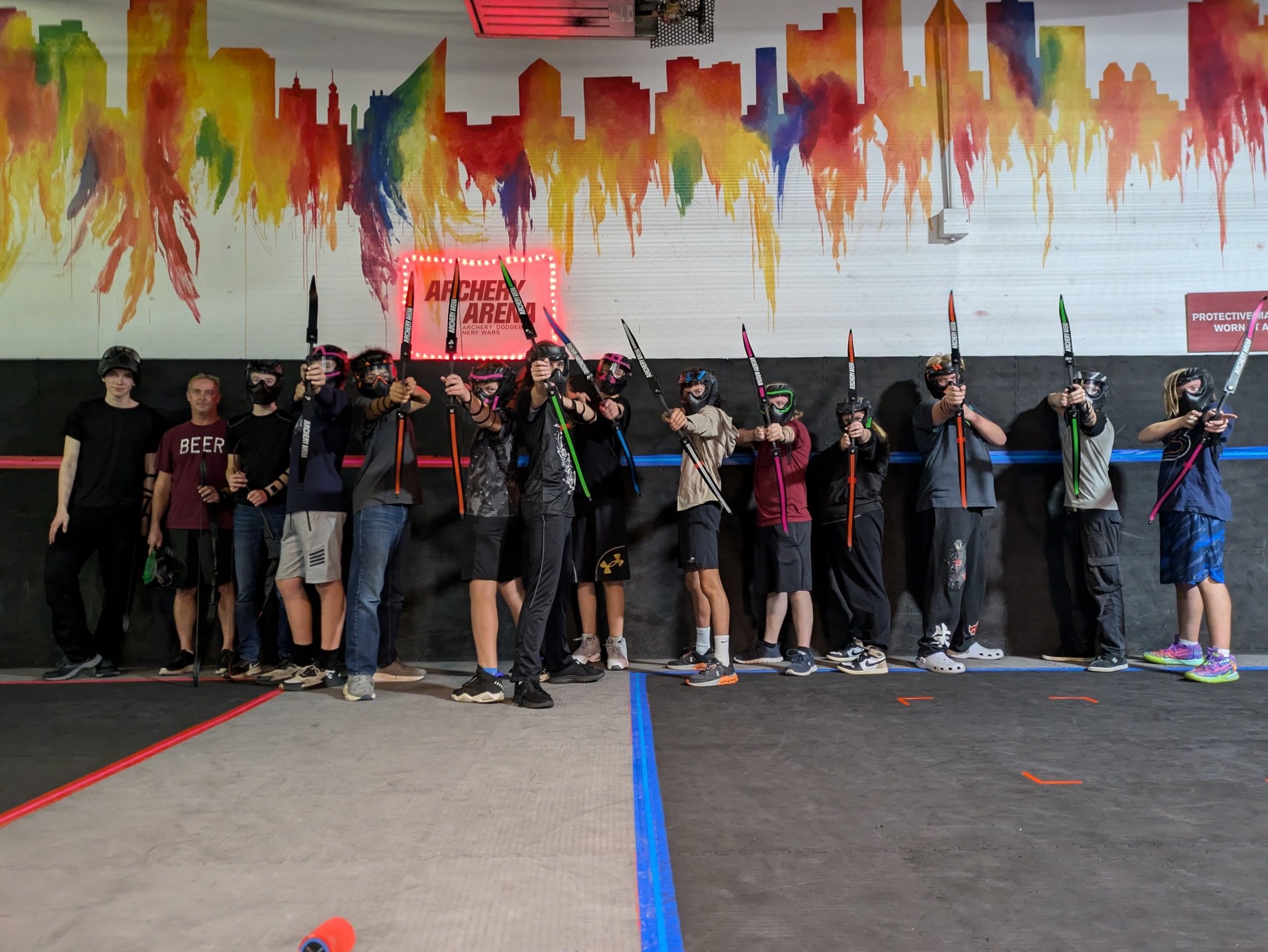 Group of children and adults at an indoor archery tag arena, holding toy bows and wearing protective gear, with colorful graffiti-style cityscape mural on the wall behind them.