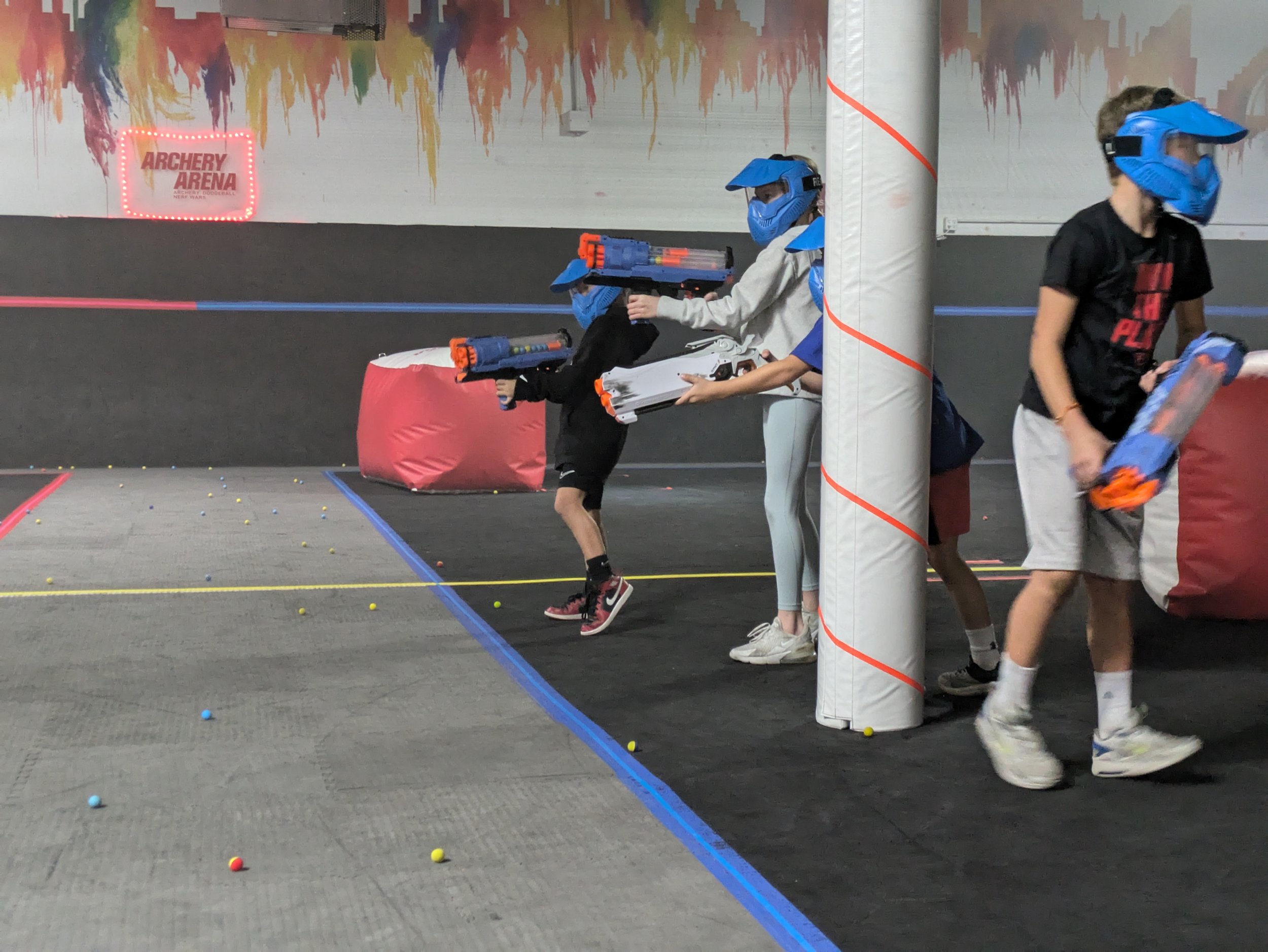 Children wearing blue helmets and holding toy laser guns while playing laser tag in an indoor arena.