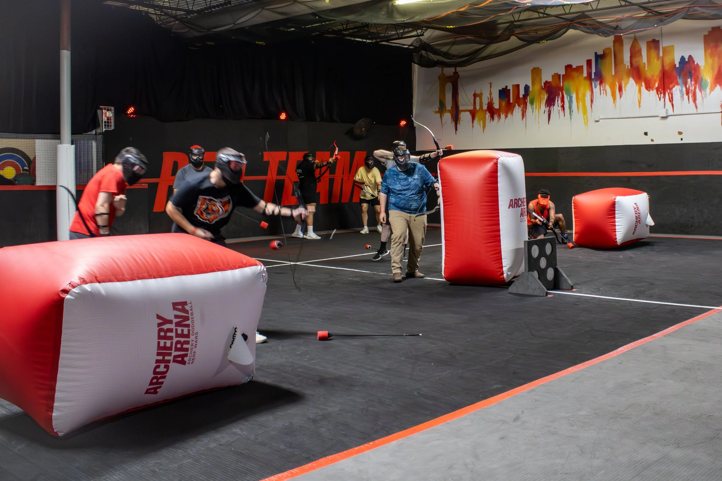 People wearing black helmets and shirts playing indoor nerf-style foam dart game, using inflatable barriers for cover in an arena with the words 'Play Time' and city skyline mural on walls.