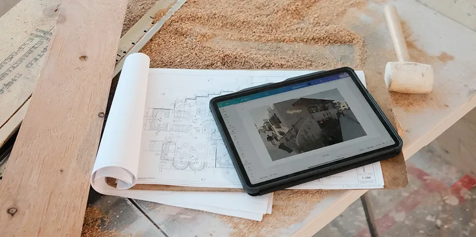 Construction drawings and a tablet showing plans on a plywood surface with sawdust at an active job site.