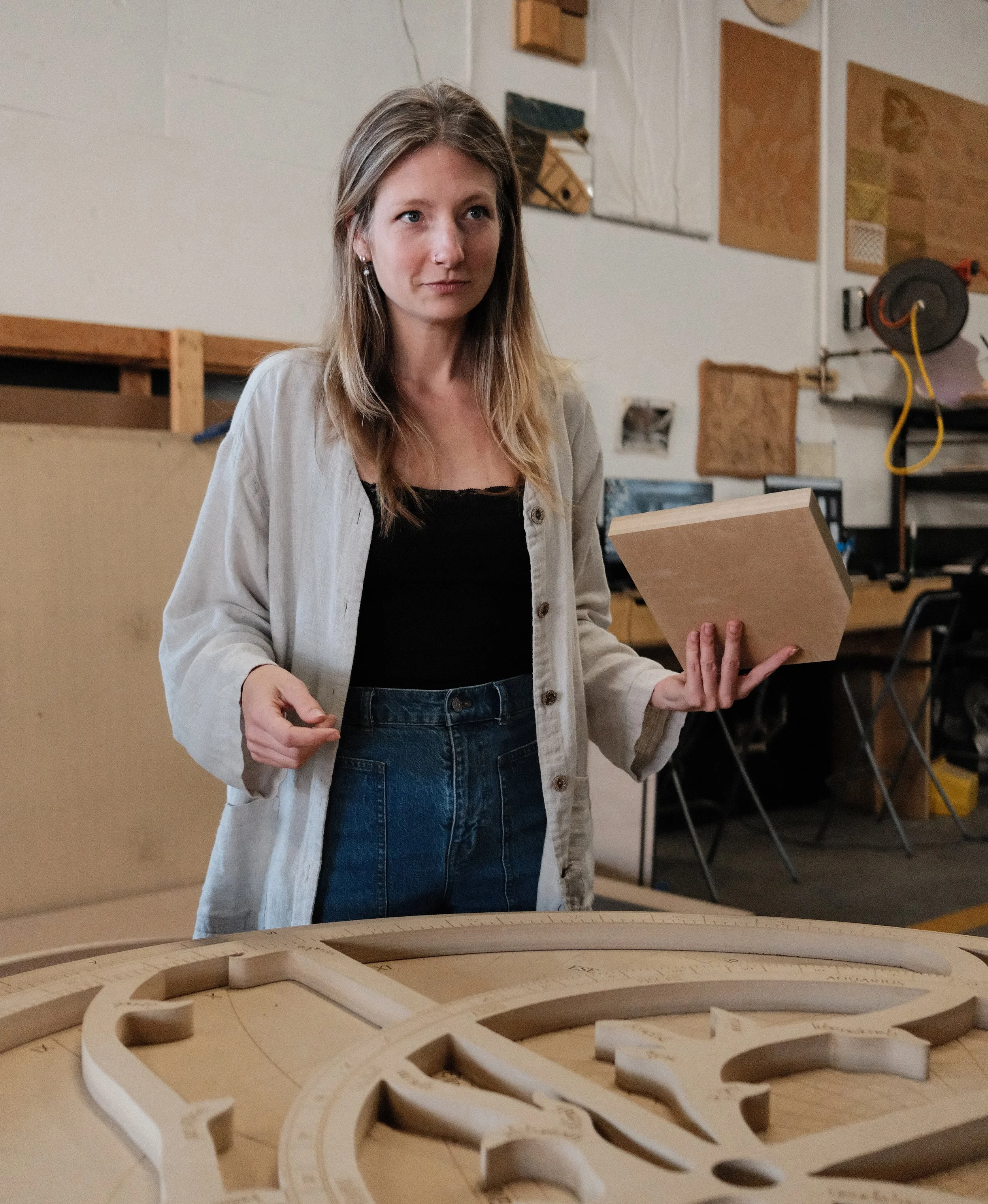 A woman with light brown hair wearing dark blue jeans, black shirt, and light grey cardigan, standing in a woodworking studio holds a wooden block while working on a large custom wood project, surrounded by tools and materials.