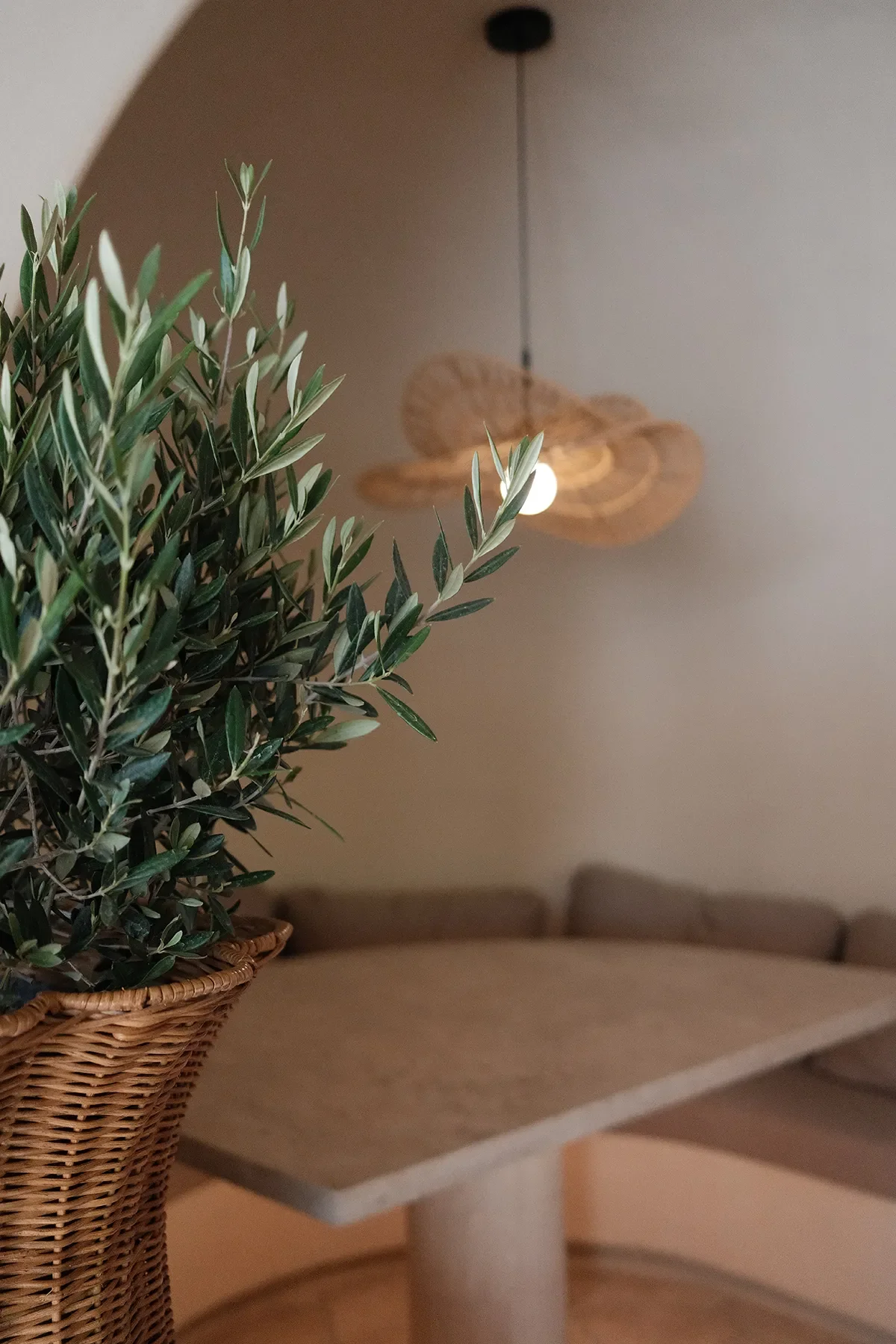 A potted tree beside a plaster table and lit rattan pendant in a booth at By The Sea