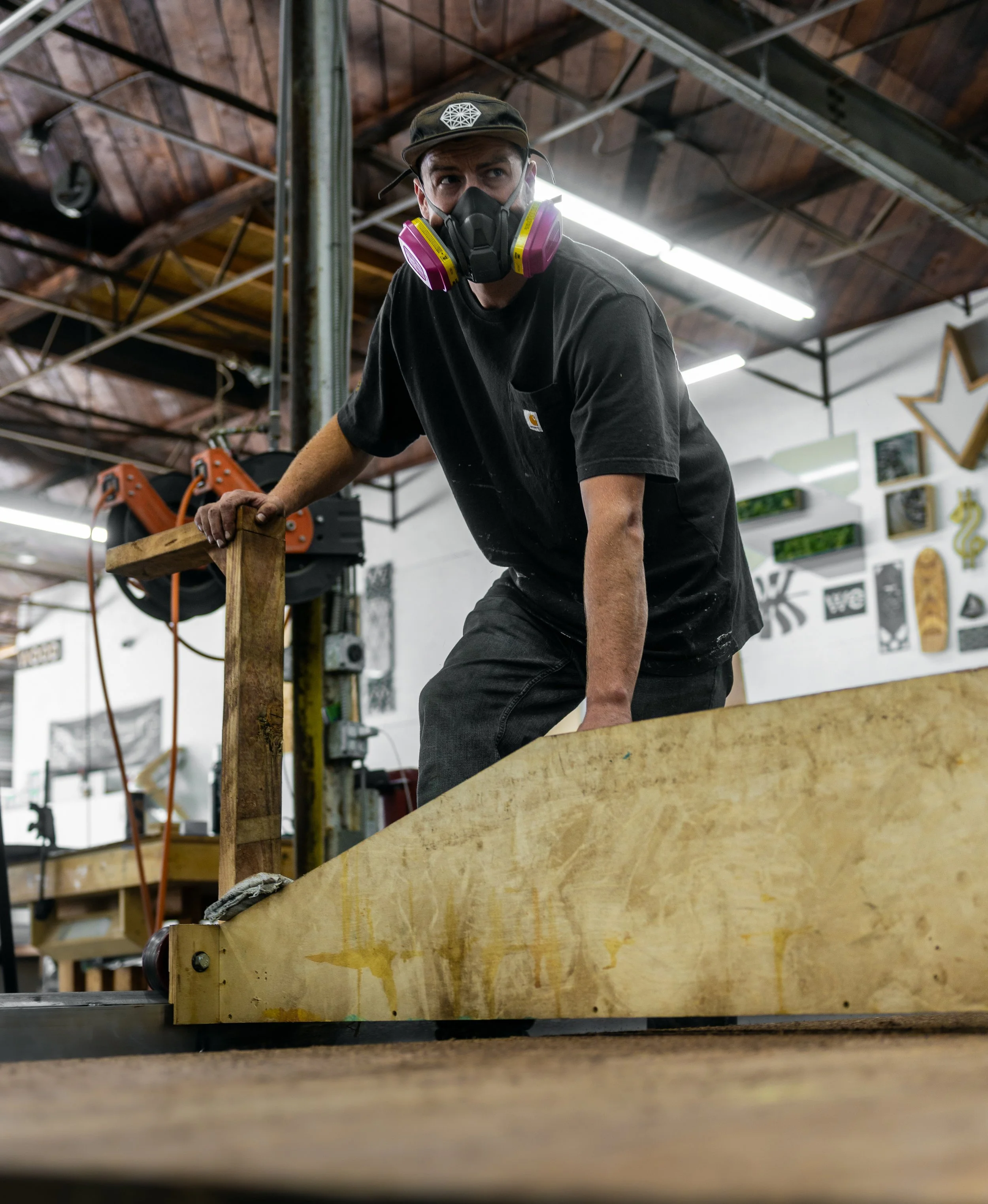 A man wearing a black t-shirt, black hat and a respirator positions a large wood component on a worktable in a workshop, preparing for fabrication.