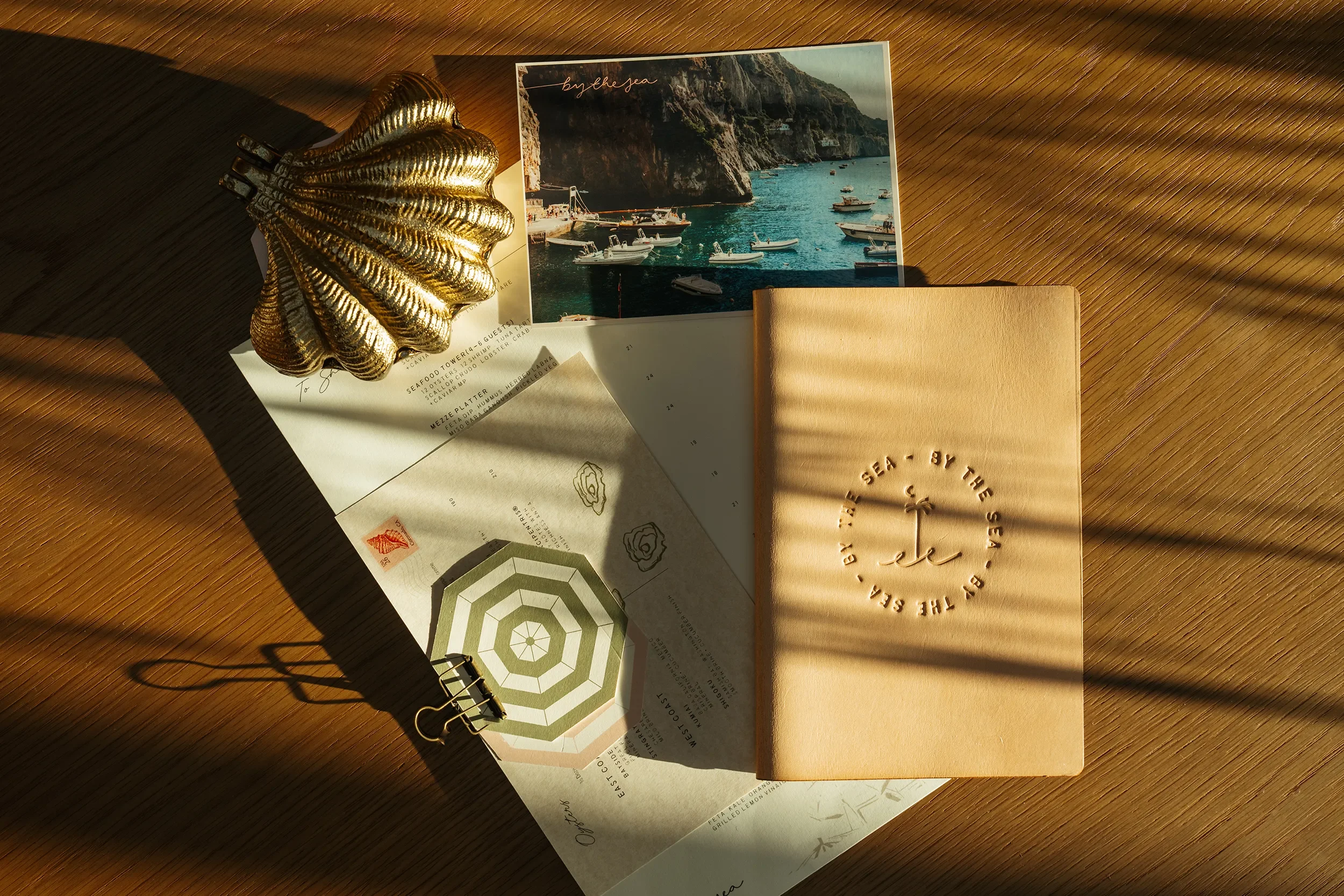 By The Sea brand collateral flat lay - embossed leather menu folio, printed menus, and a brass shell on oak wood
