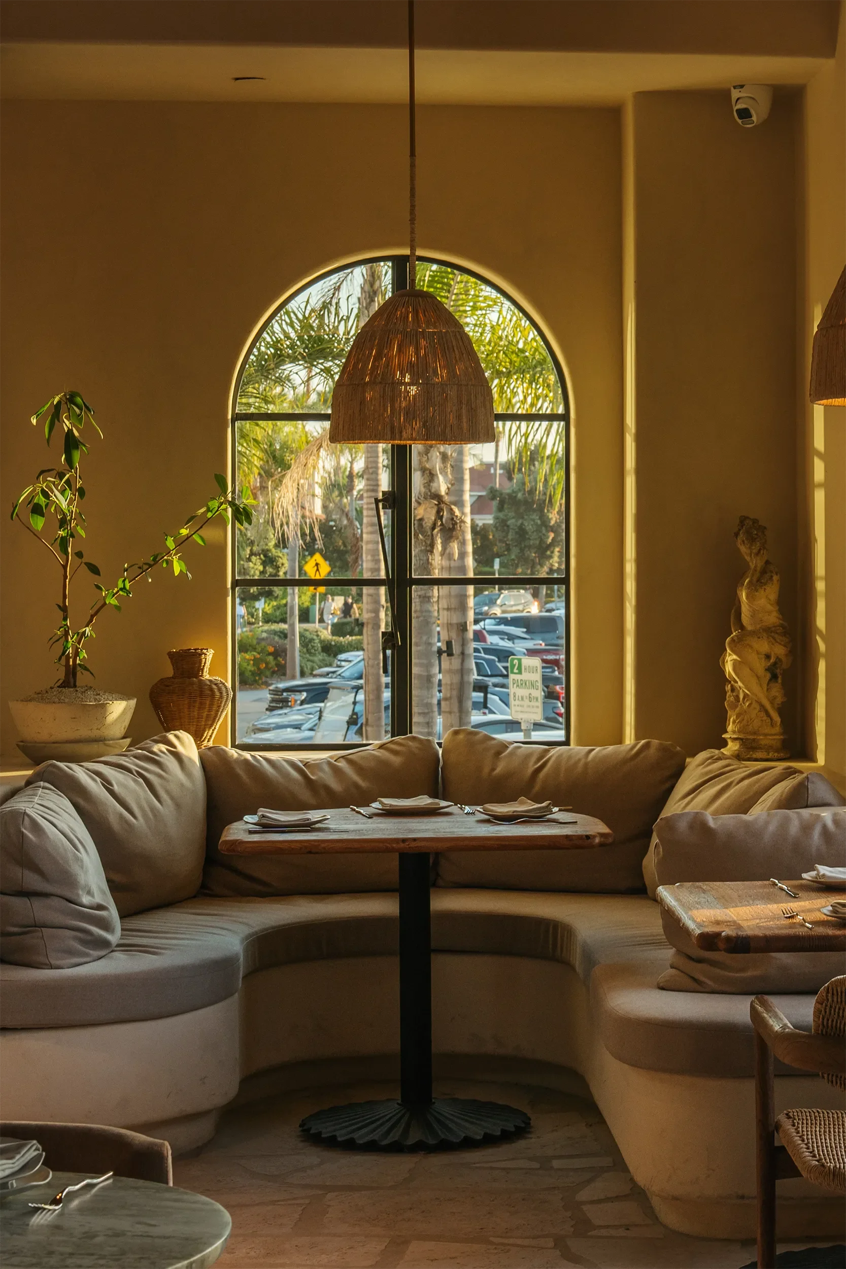 Corner banquette under a tall arched window and rattan pendant in the front dining room at By The Sea