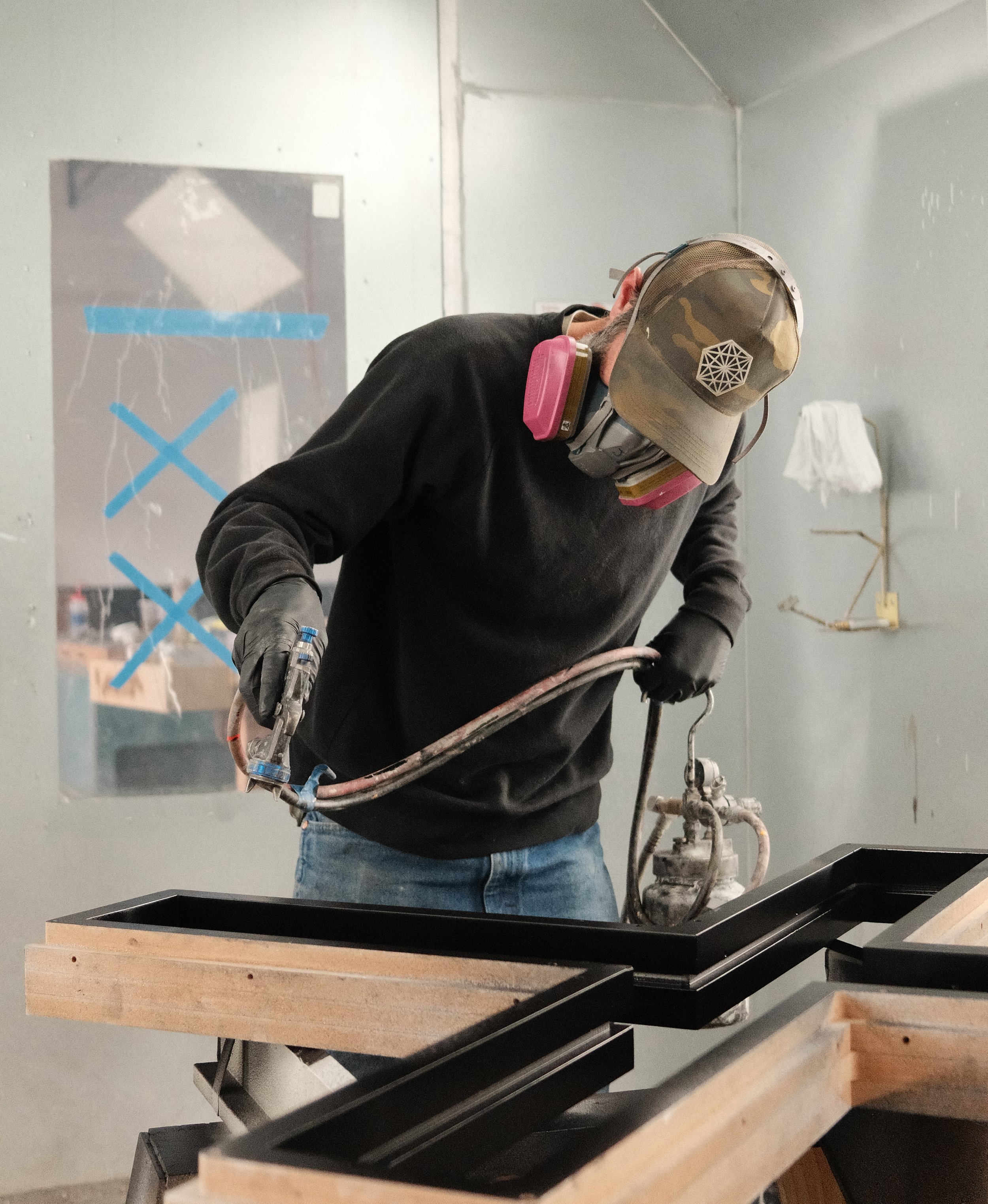 A man wearing a black sweatshirt, blue jeans, a hat and a respirator uses a spray gun to apply finish to custom wood components in a workshop spray booth.