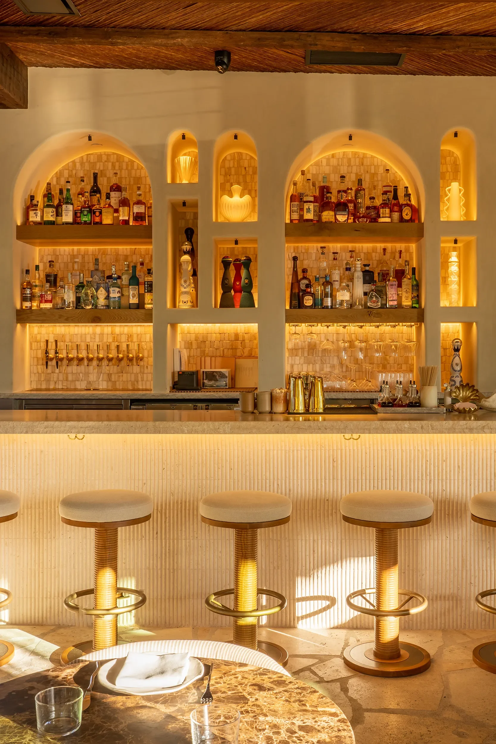 Backlit arched liquor niches and upholstered stools along the travertine bar at By The Sea