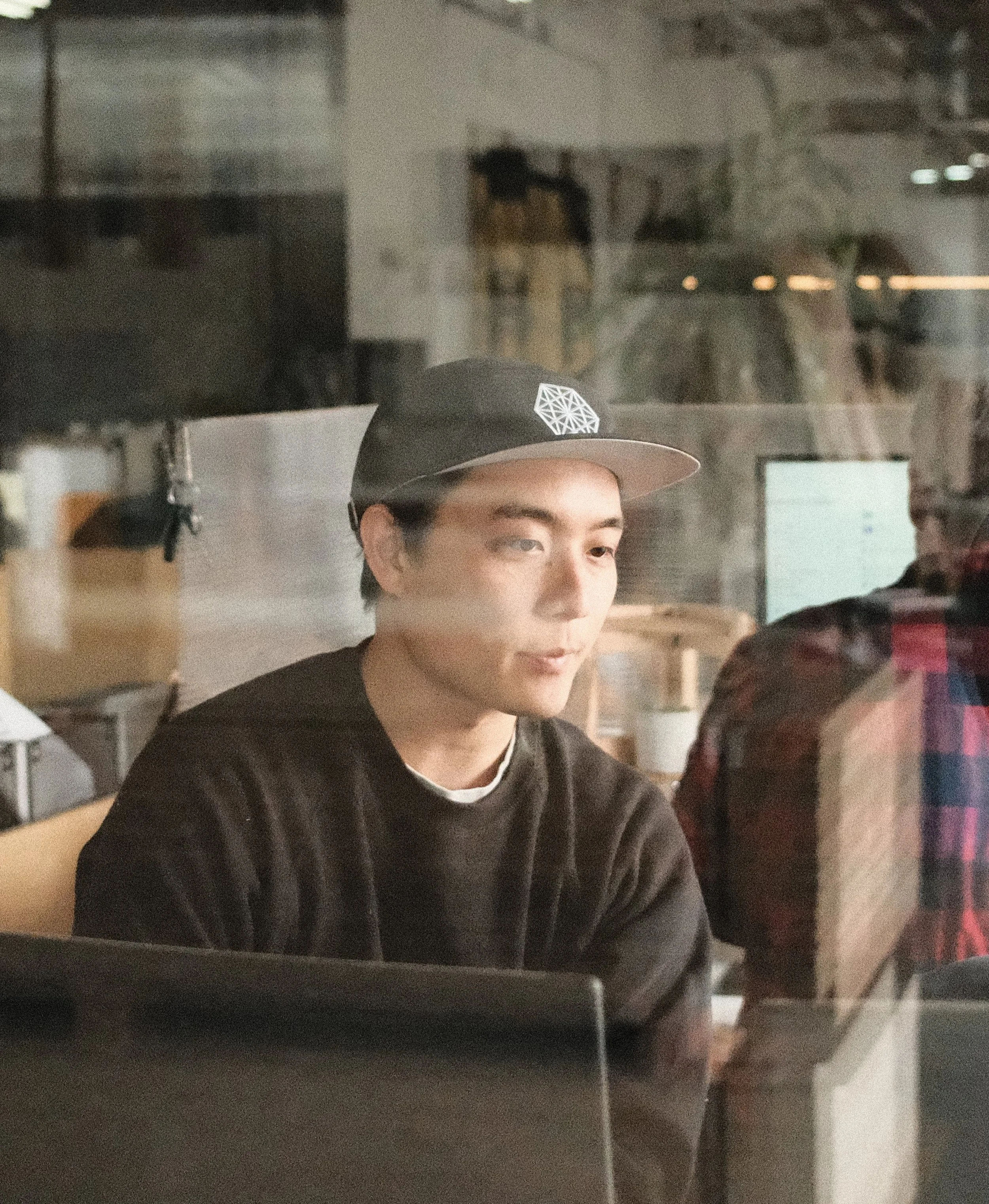 A man wearing black sweatshirt and black hat, sitting behind glass with reflections of the surrounding office, works on a laptop in a studio office, viewed through glass, focused on design and project planning.