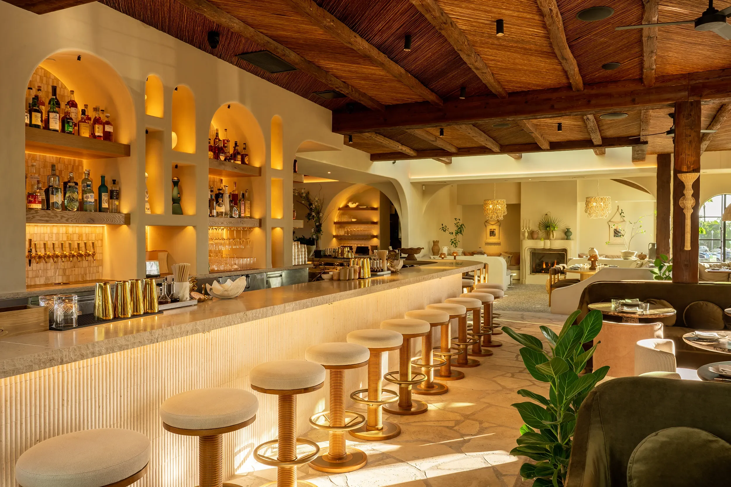 Curved travertine bar at By The Sea with a fluted front, backlit arched liquor niches, and a beamed ceiling