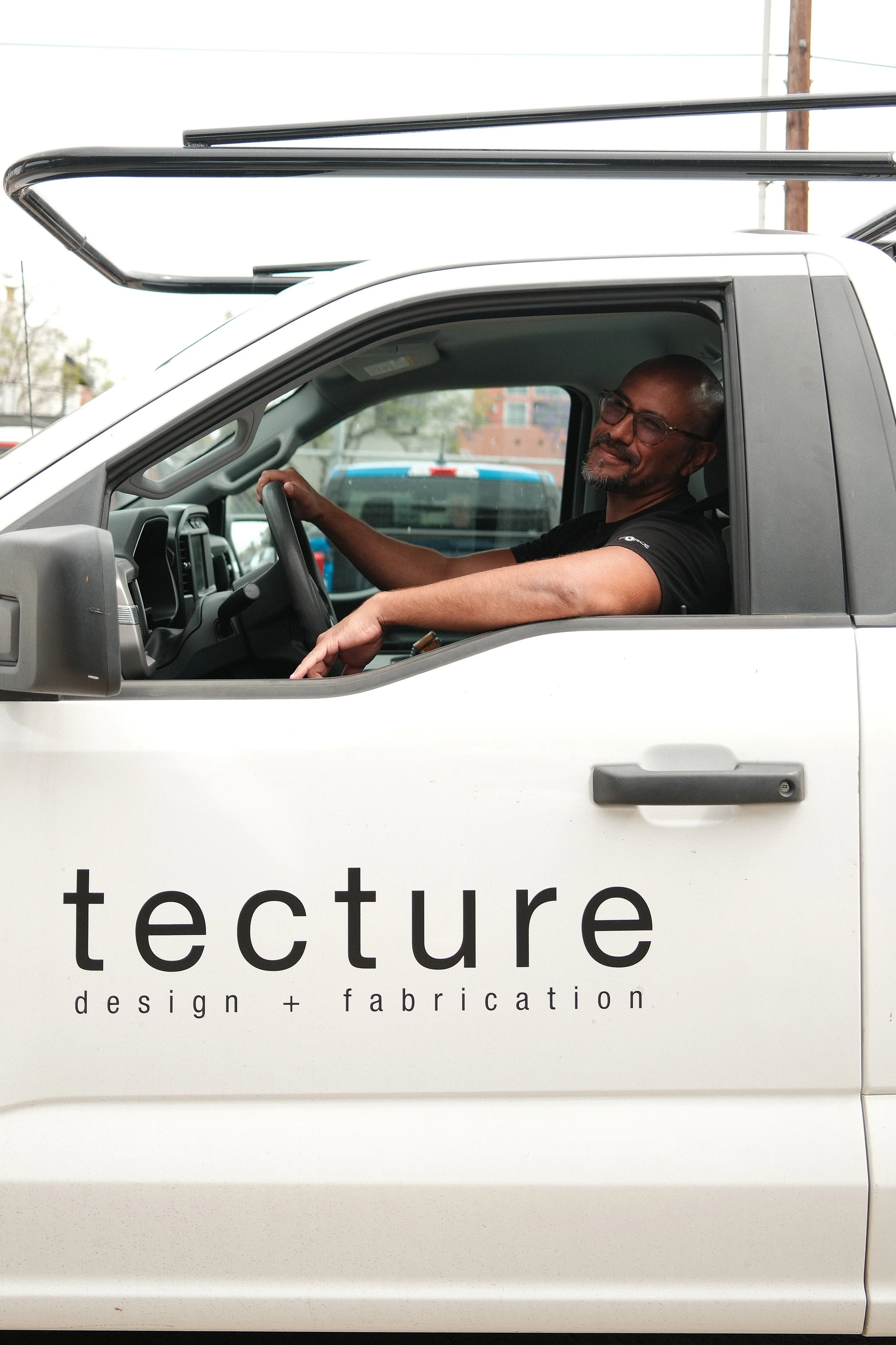 A man with a shaved head and a gray goatee, wearing glasses and a black short sleeve t-shirt, sits in the driver’s seat of a branded white work truck, highlighting design and fabrication services on-site.