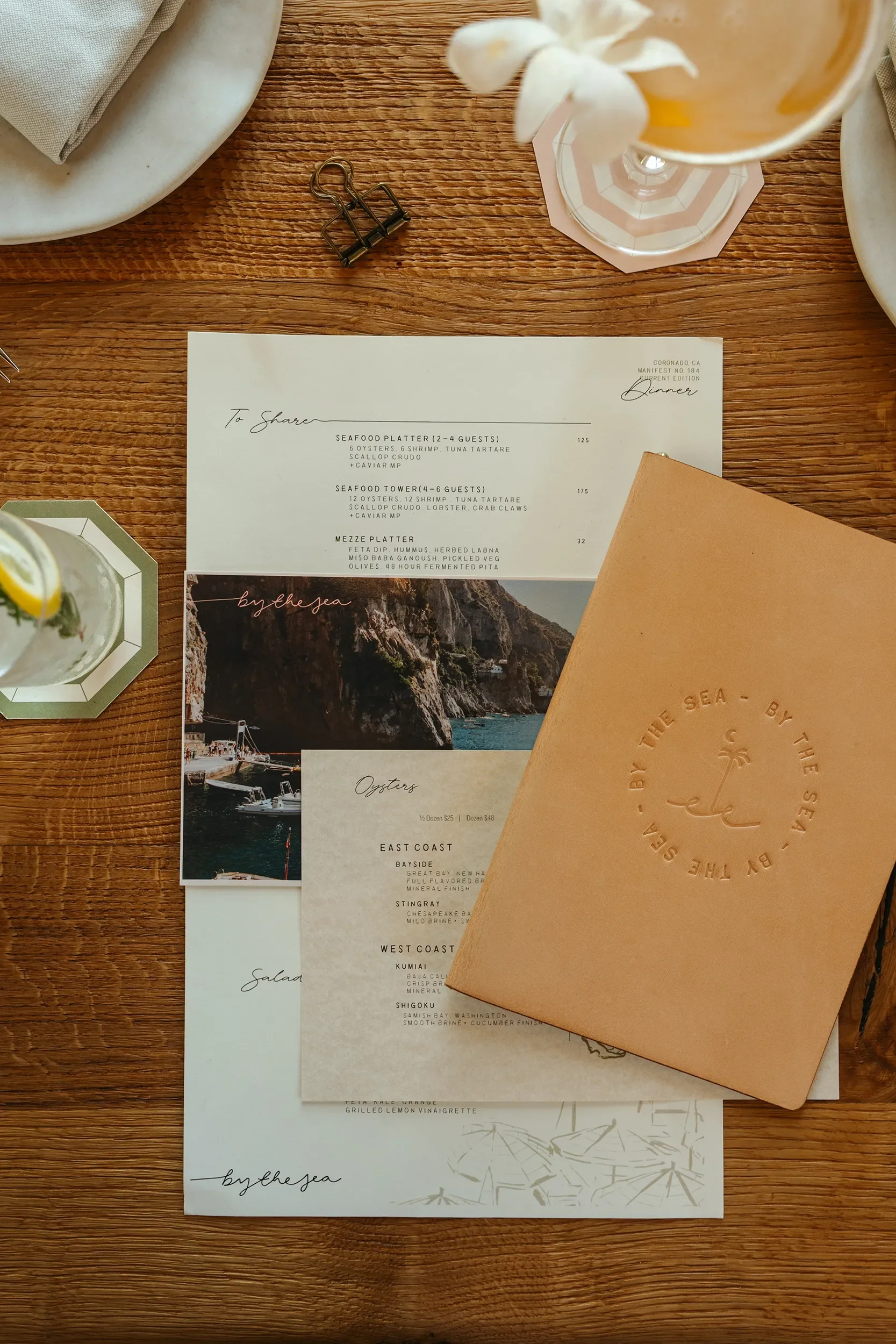Debossed leather menu folio and printed food menus styled with a cocktail and coastal postcard at By The Sea