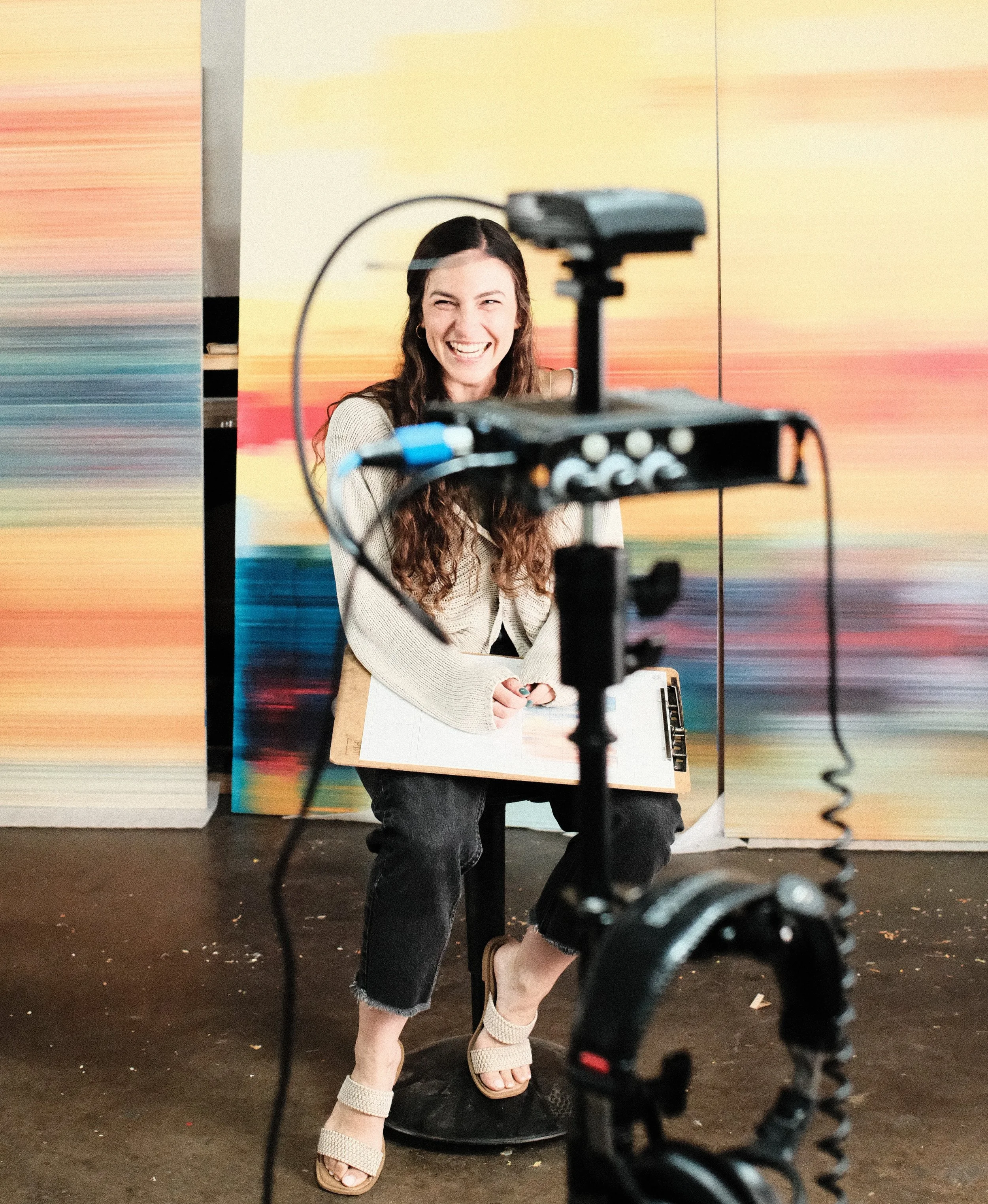 A woman with long brown curly hair wearing black jeans and a beige sweater, sits in a studio in front of colorful custom abstract paintings, laughing while being filmed on camera equipment.