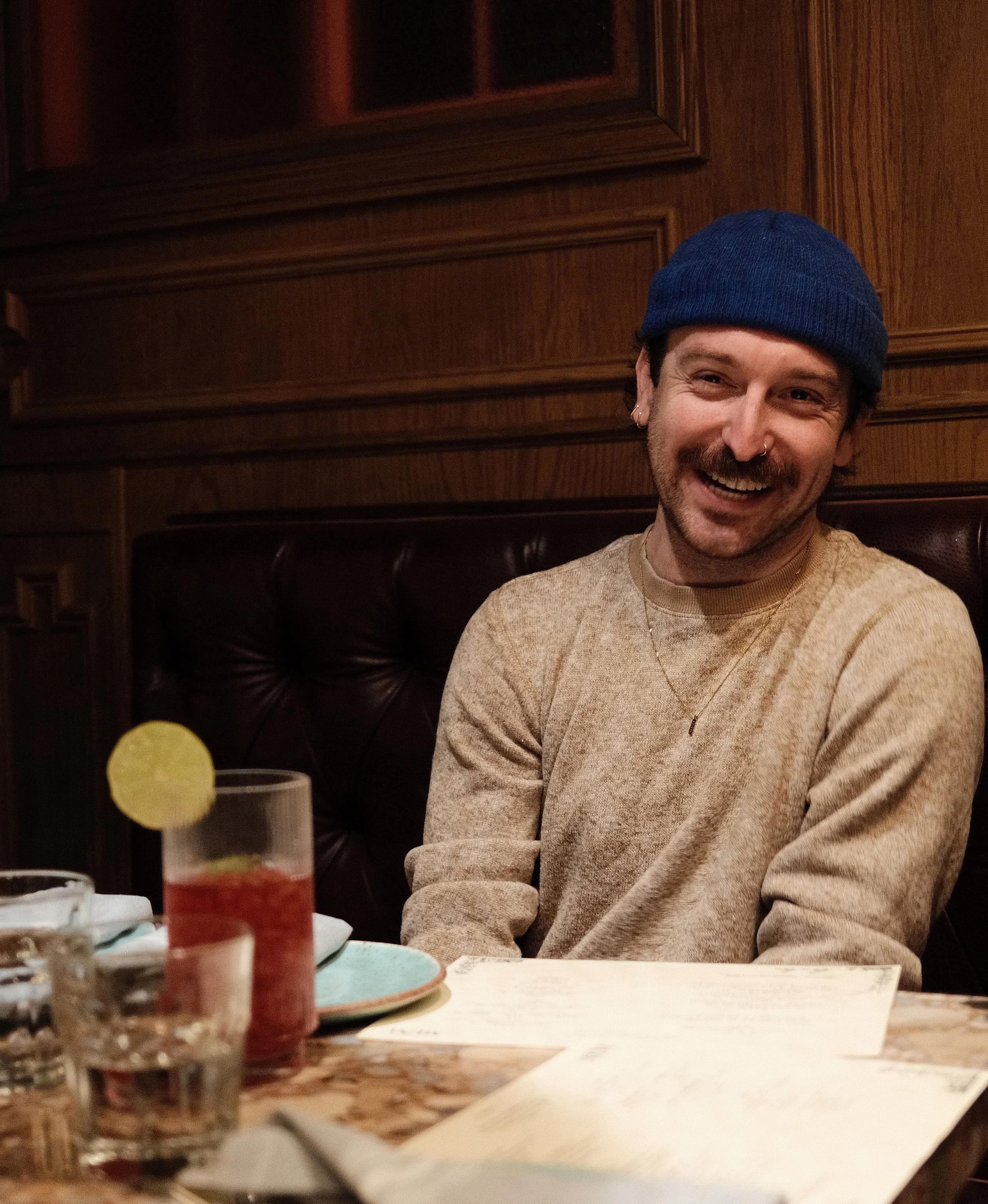 A smiling man with a mustache, wearing a beige sweatshirt and a blue beanie sits at a restaurant table with drinks and menus, enjoying a casual dining moment.