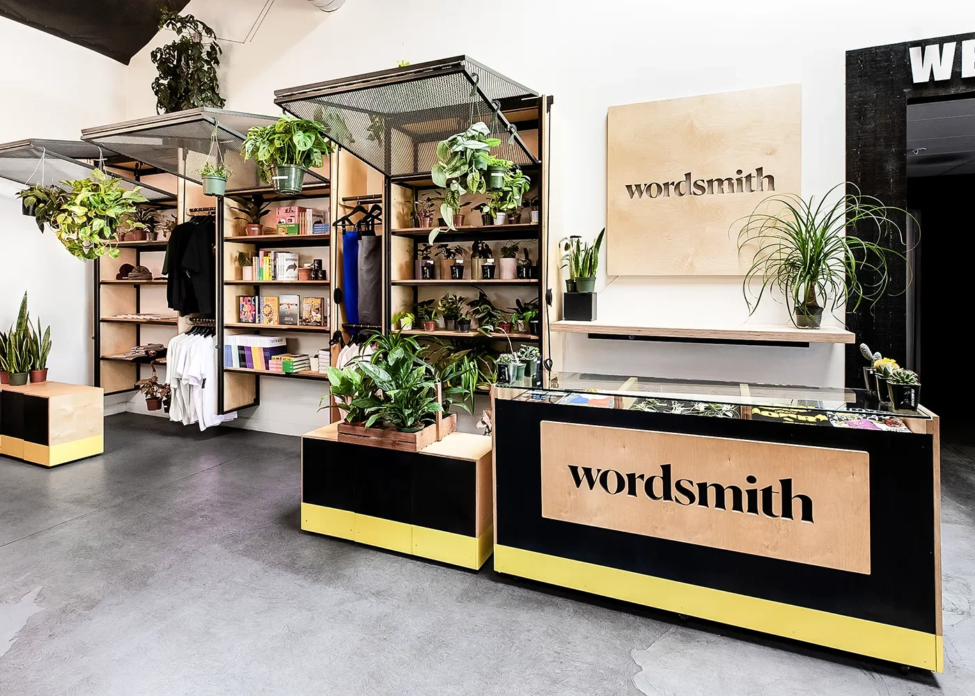Wordsmith retail interior with a branded checkout counter, wood and black metal shelving, hanging plants, and yellow accents.