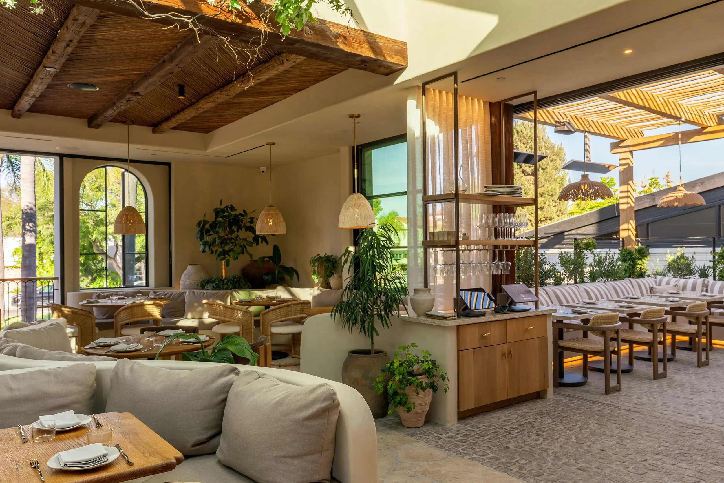 Indoor-outdoor lounge at By The Sea with arched doors, rattan pendants, and a view to the pergola terrace