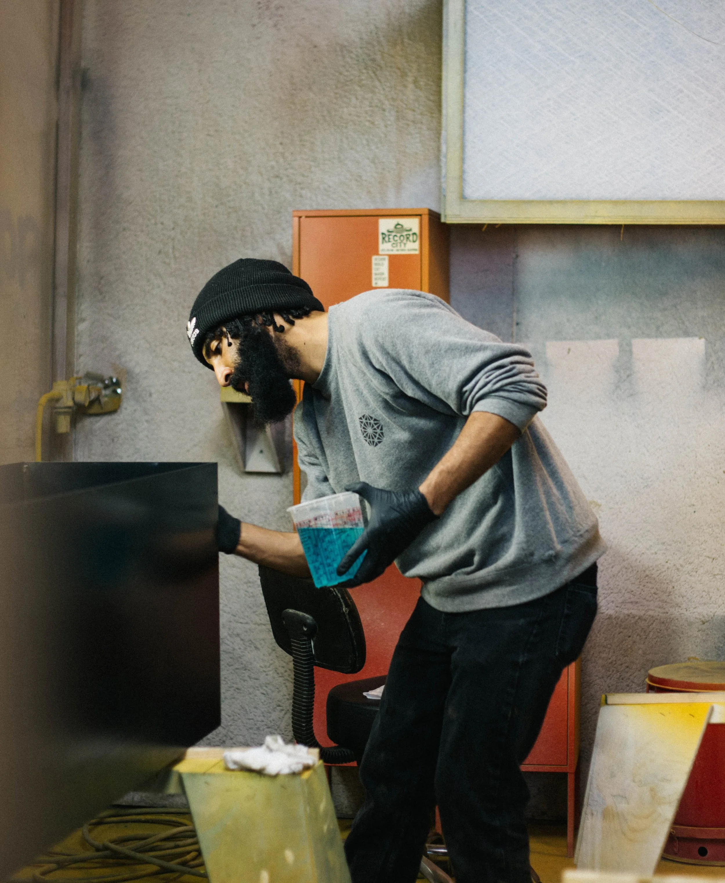 A man with a long dark beard wearing a gray sweatshirt, black pants and black beanie, in a workshop mixes and pours finishing liquid onto a surface, focused on detail work in a production setting.