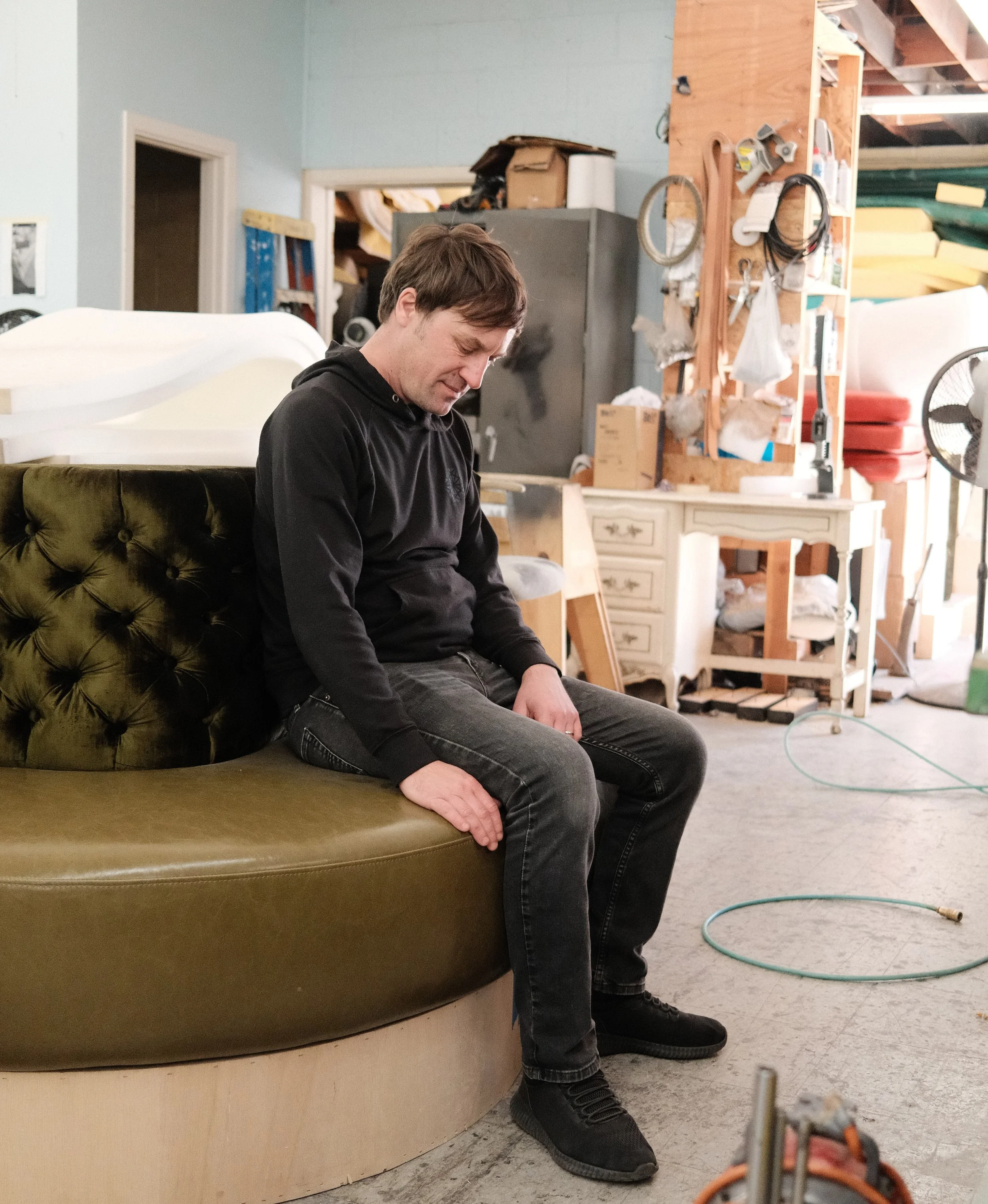 A man with light brown hair in black clothing sits on a custom curved olive green upholstered banquette with leather seat and tufted velvet back and light wood base, in a workshop, inspecting craftsmanship details.