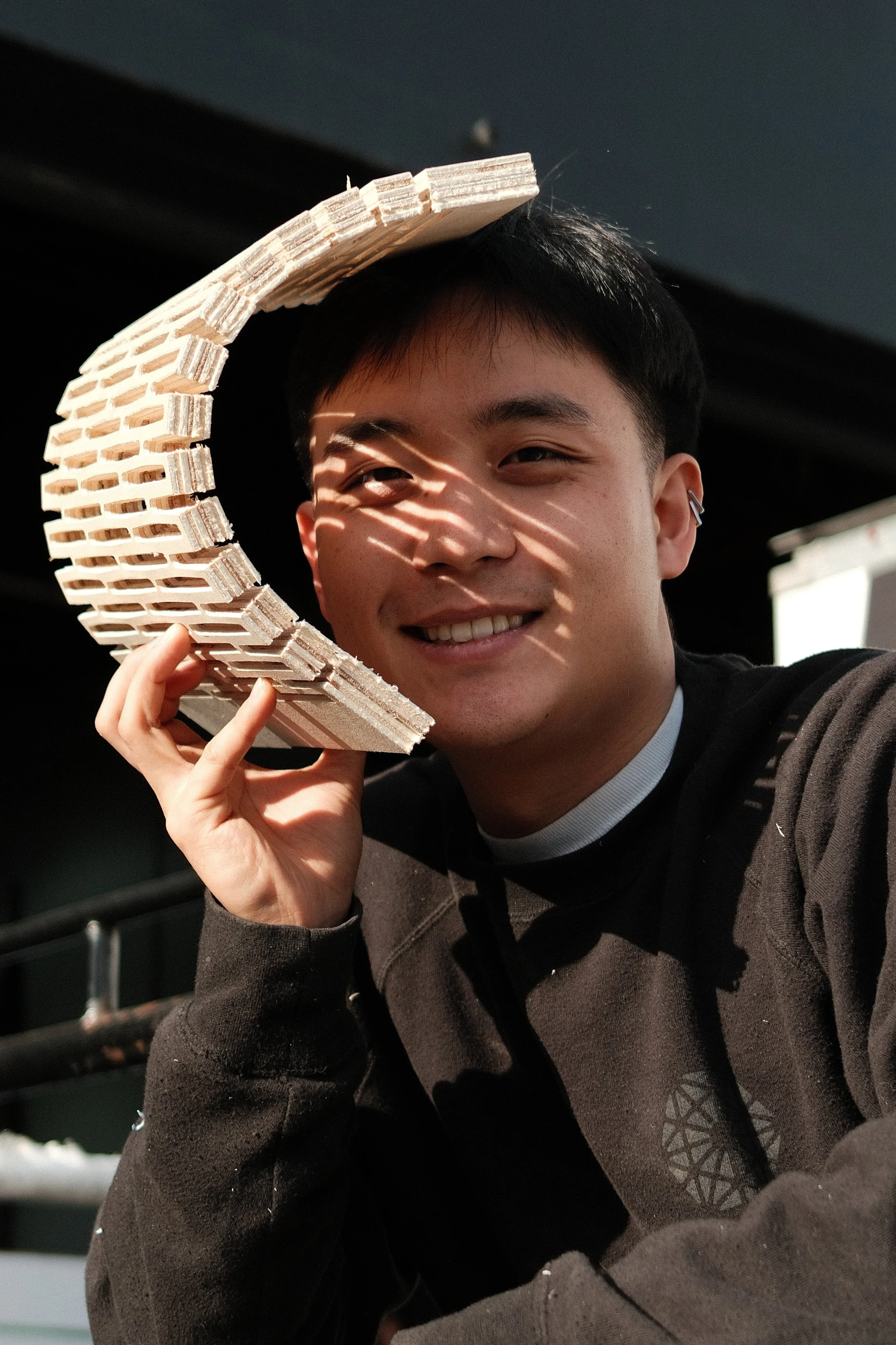 A man wearing black sweatshirt and black hat, sitting behind glass with reflections of the surrounding office, works on a laptop in a studio office, viewed through glass, focused on design and project planning.