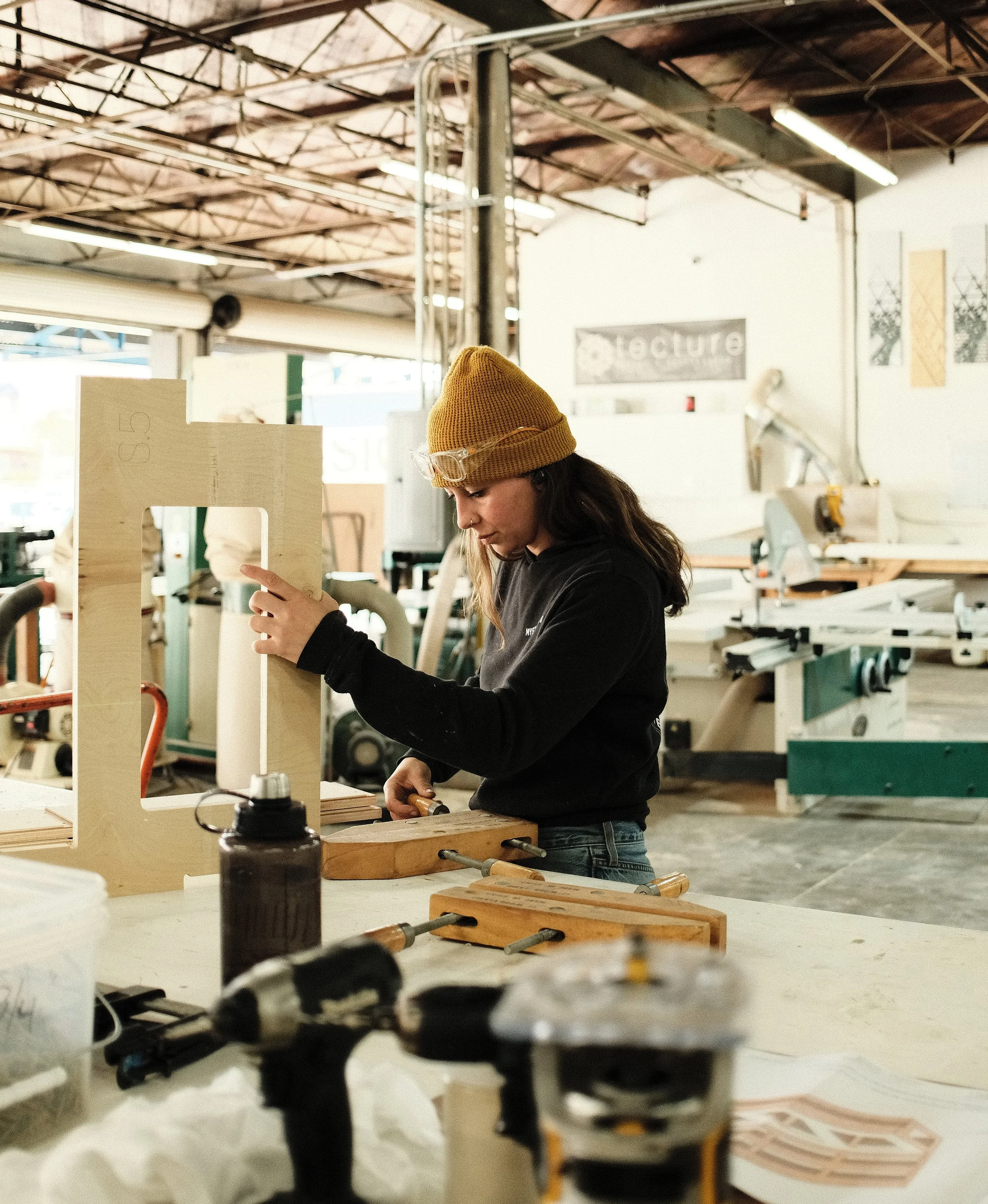 A woman with medium length brown hair wearing a yellow beanie and black sweatshirt, working in a woodworking shop carefully assembles a custom wood component using clamps and tools at a workbench.