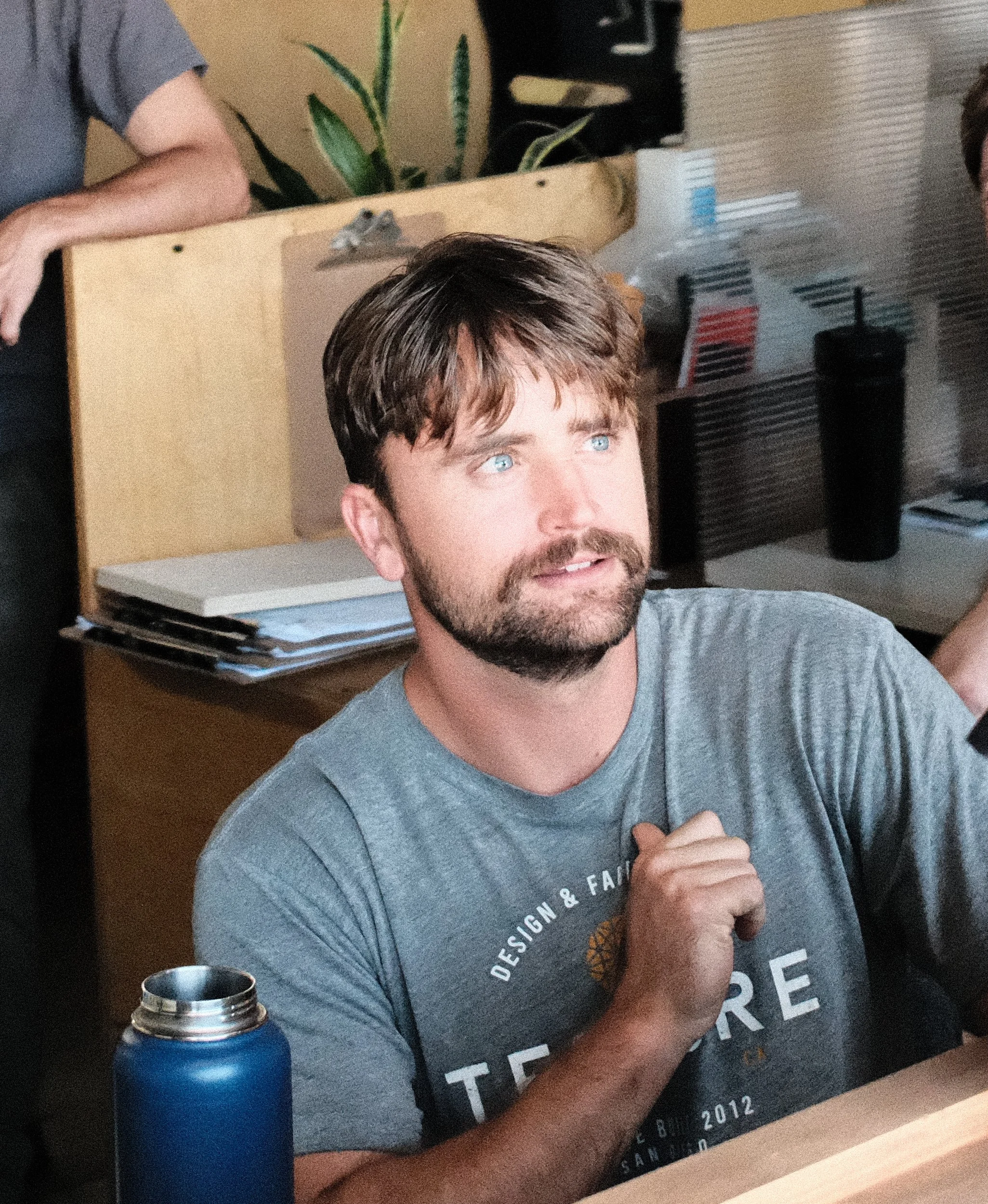 A man with brown hair and facial hair with blue eyes, wearing a gray Tecture design and fabrication t-shirt sits in an office workspace, engaged in conversation beside a wooden desk with tools and materials.