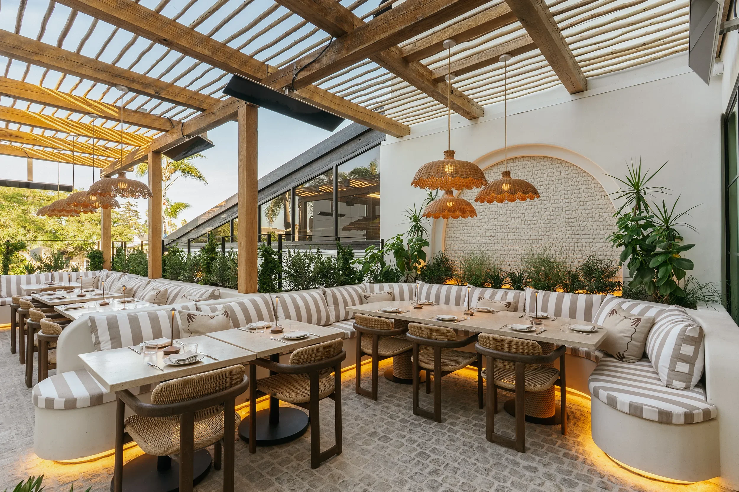 Open-air terrace at By The Sea with eucalyptus trellis, rattan pendant lighting, and striped banquette seating