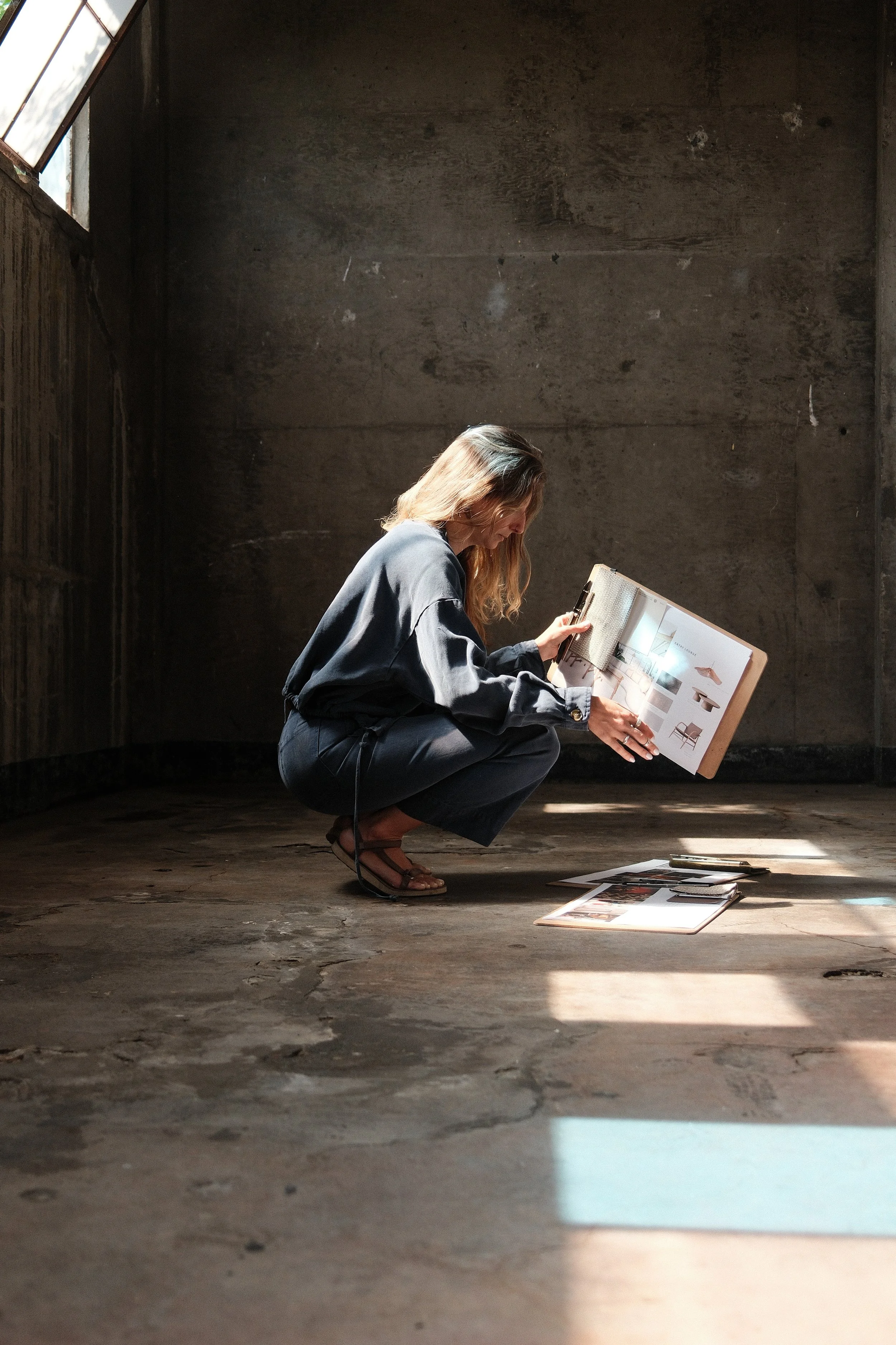 A woman with light brown hair wearing dark blue jeans, black shirt, and light grey cardigan, standing in a woodworking studio holds a wooden block while working on a large custom wood project, surrounded by tools and materials.