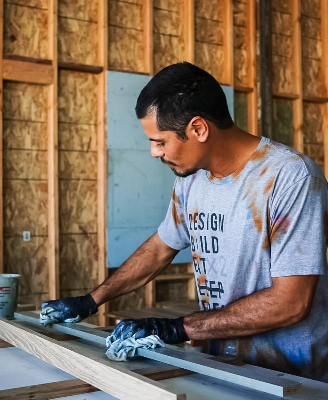 A man in a light gray Tecture t-shirt with brown paint marks scattered on it, in a workshop wipes down a painted wood component, focusing on finishing details and surface quality.
