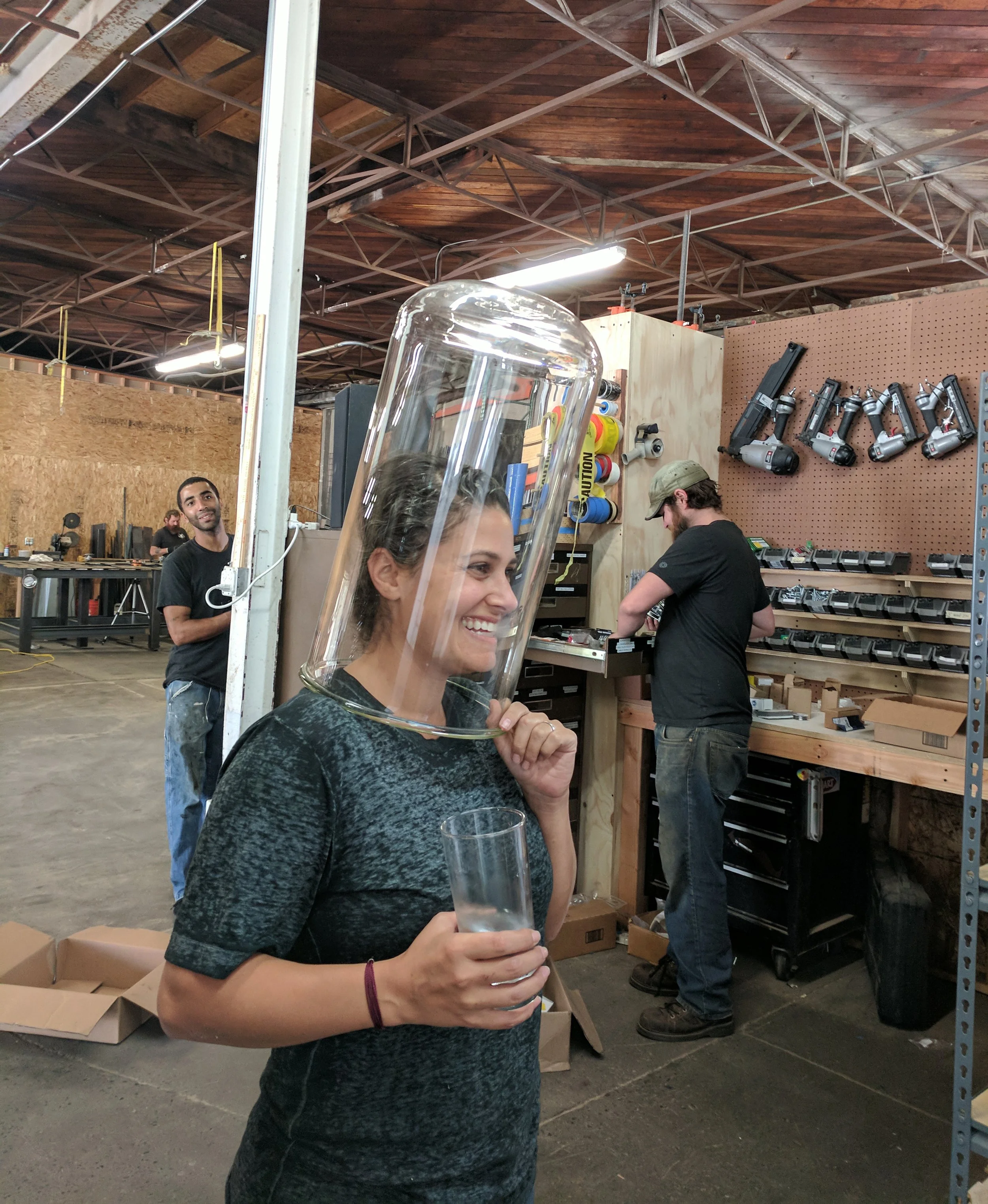 A woman in a workshop smiles while holding a drinking glass and playfully wearing an oversized glass jar over her head, with coworkers using tools in the background.