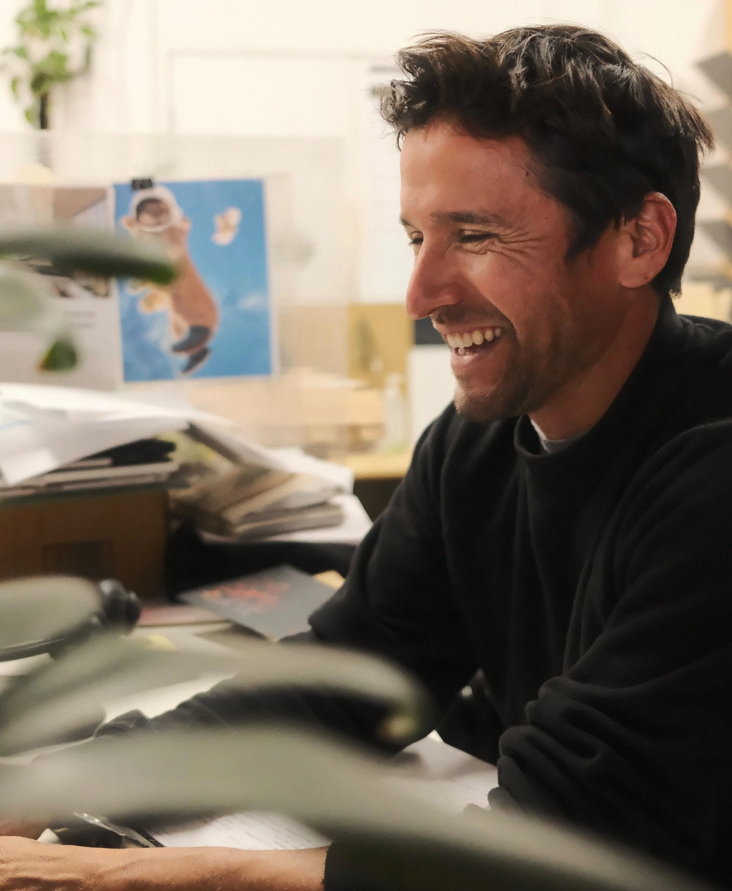 A man with dark brown hair , wearing a black long sleeve shirt smiling and laughing at his desk on his computer.