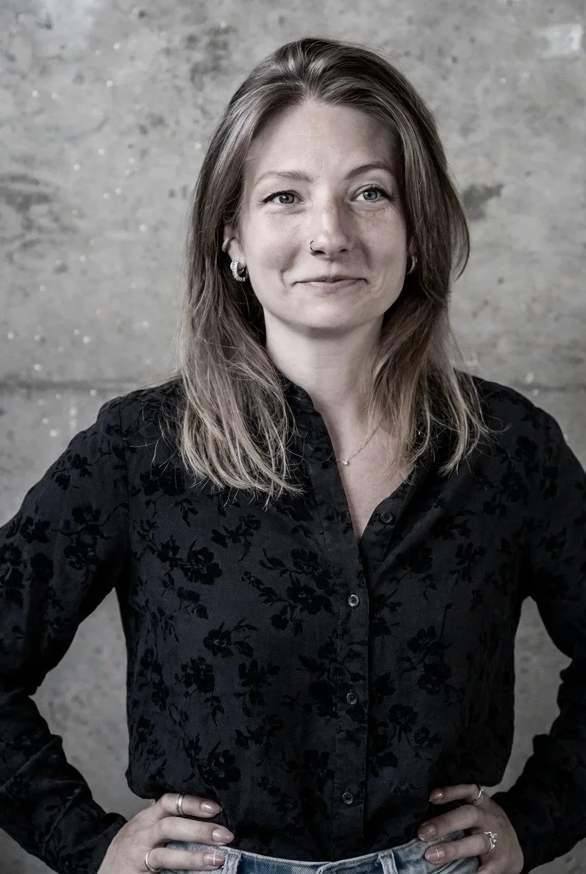 A woman with shoulder-length light brown hair, wearing a patterned blouse and jeans, smiling with her hand on her hip, standing against a concrete wall.