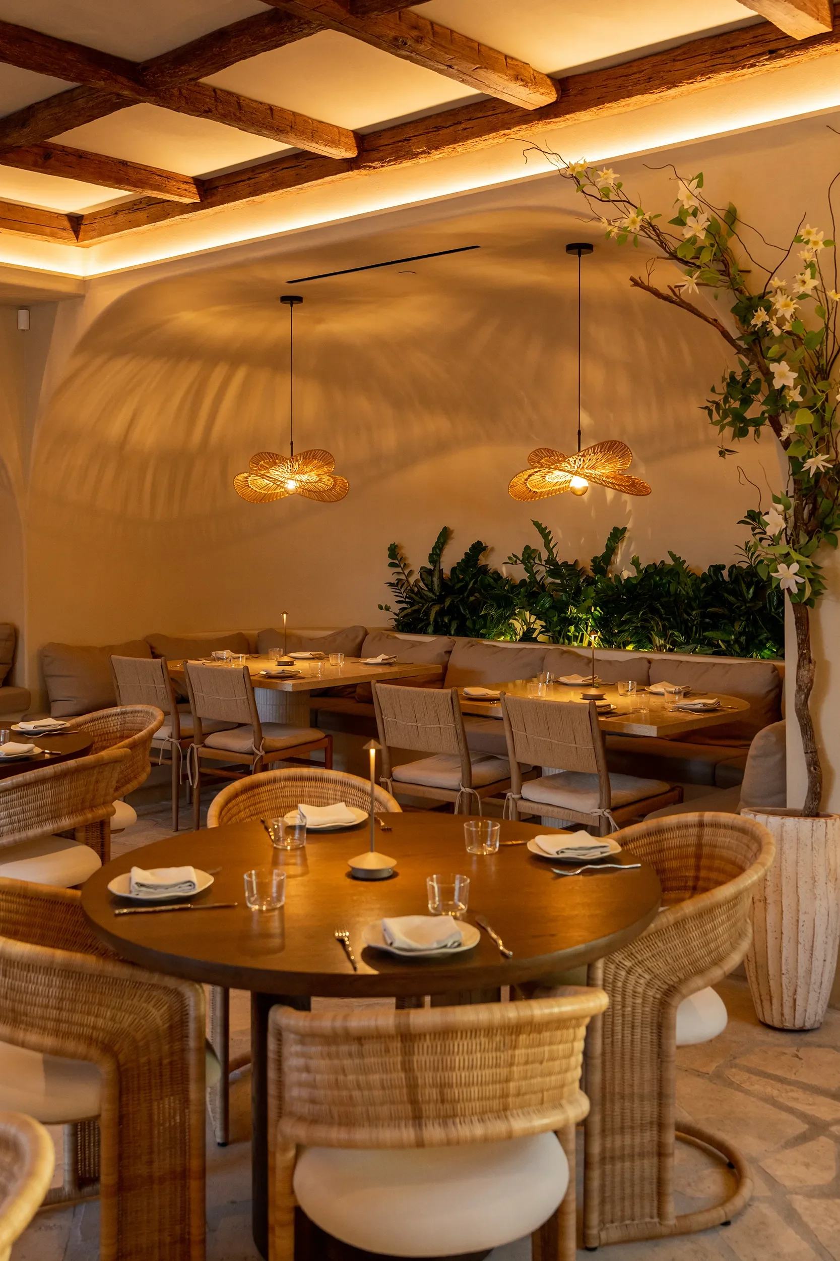 Round walnut tables and rattan chairs beneath cove-lit beams and blossom branches at By The Sea