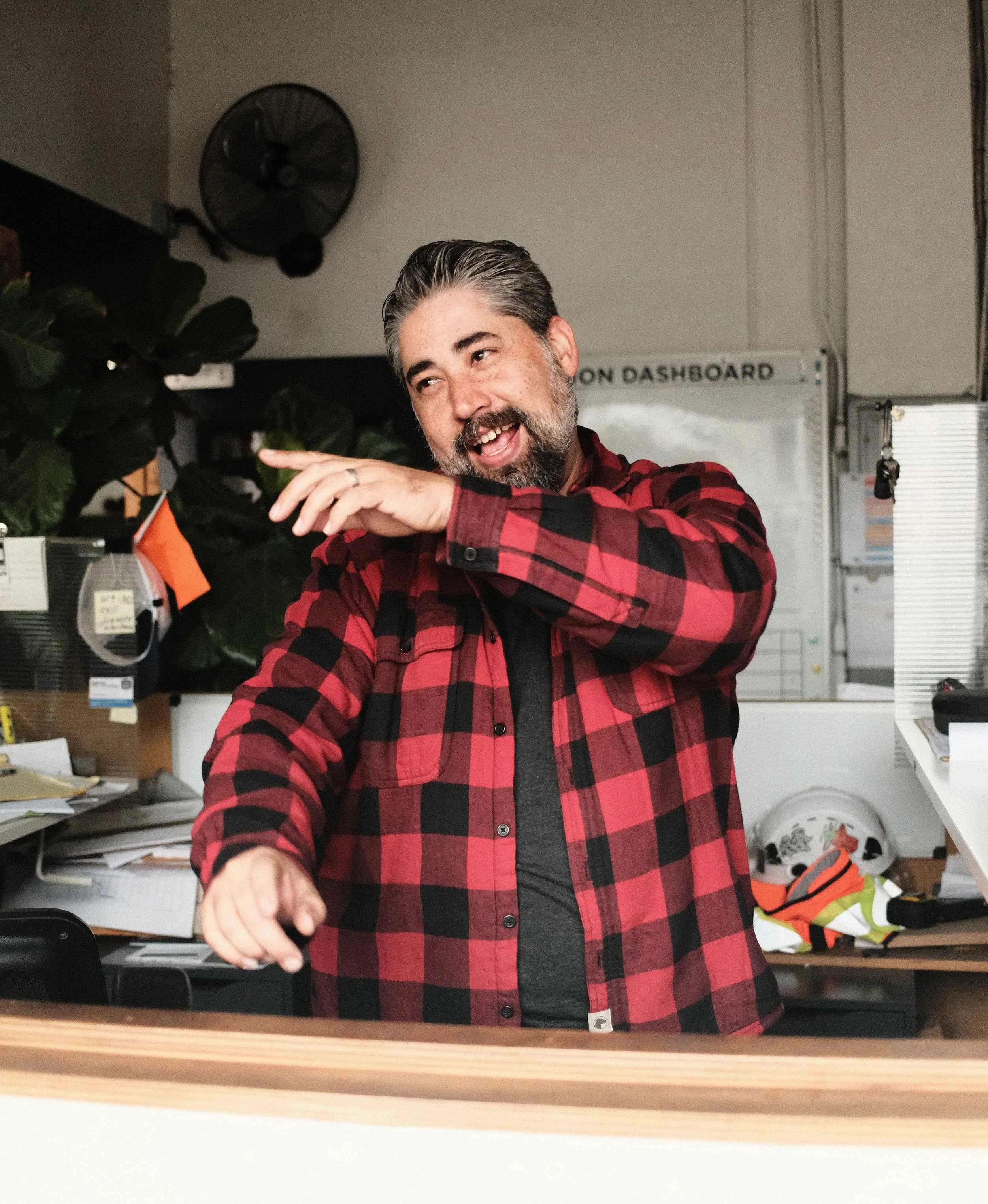 A man with a beard and gray hair, smiling and gestures mid-conversation inside a workshop office, with construction plans, tools, and safety gear visible in the background.