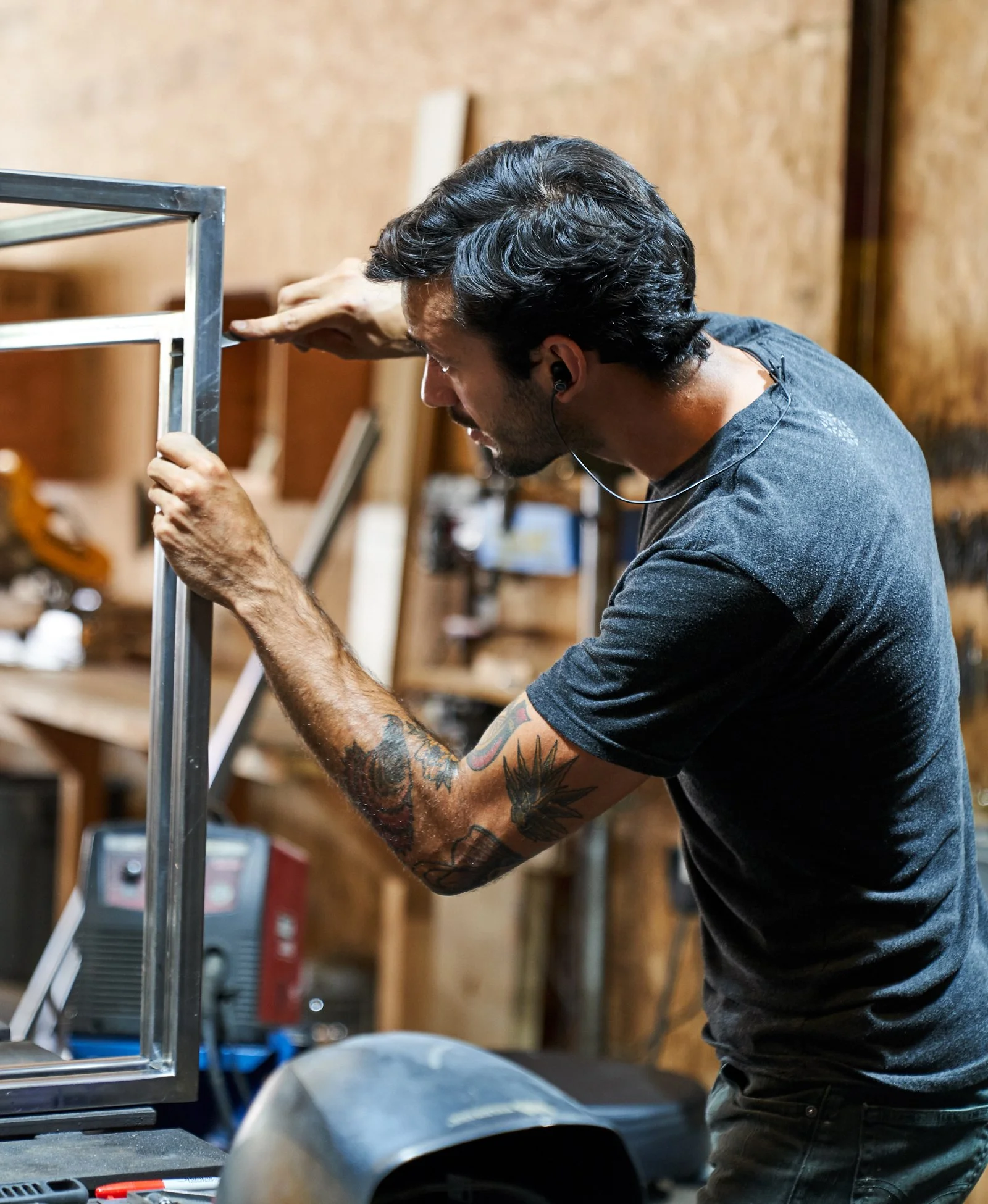 A man with dark brown hair wearing a grey t-shirt and has tattooed arms carefully aligns a steel frame in a workshop, using precision tools and equipment surrounded by industrial materials.